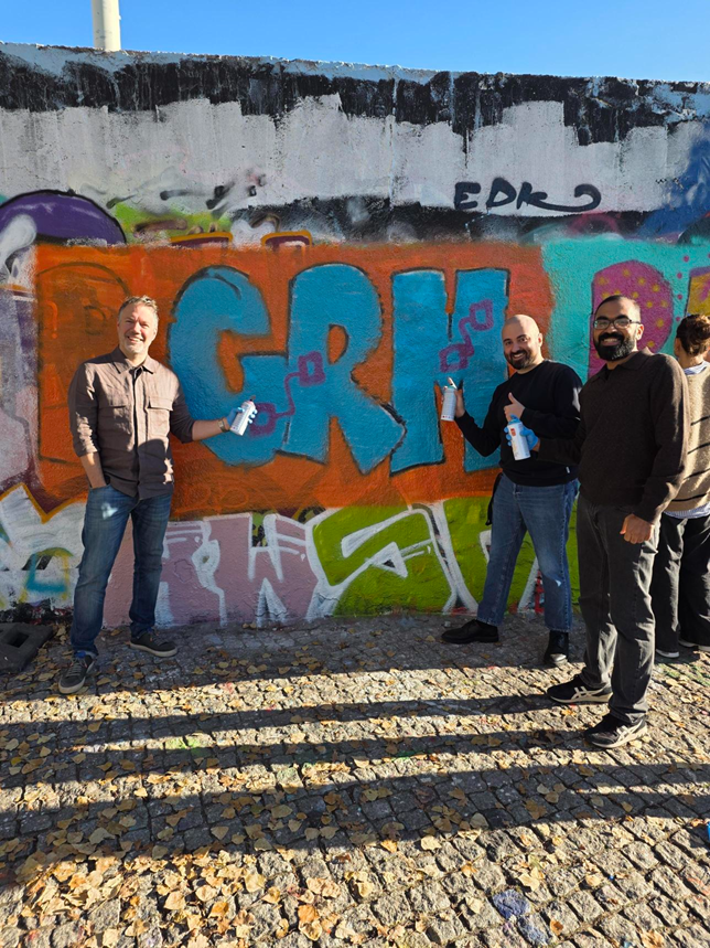 Three men standing in front of a colorful graffiti wall outdoors, smiling and holding spray paint cans.