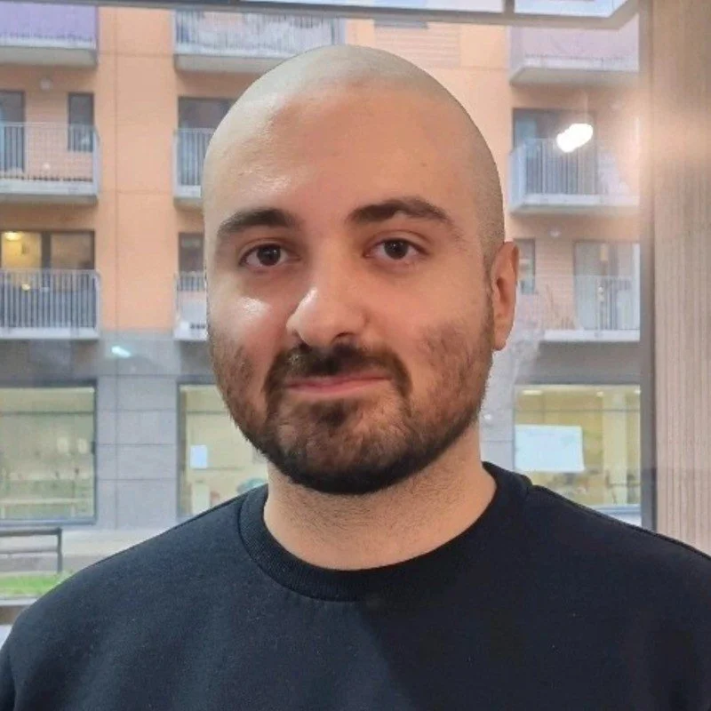 Portrait of a smiling Asian woman with shoulder-length black hair, wearing a blue top, standing outdoors with green foliage in the background.