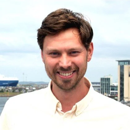 Black and white portrait of a smiling man with a thick beard, shaved head, and light-colored eyes, wearing a black shirt.