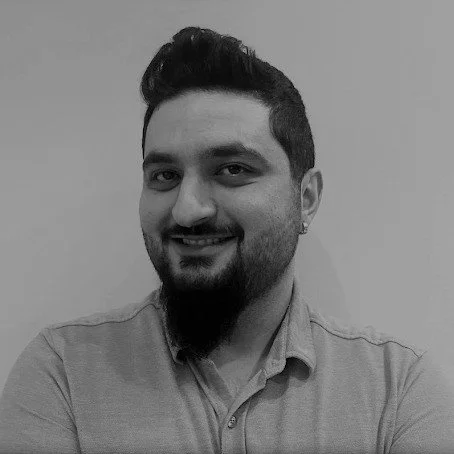 A young man with dark hair, fair skin, and hazel eyes, wearing a grey sweater over a collared shirt, smiling slightly against a neutral background.