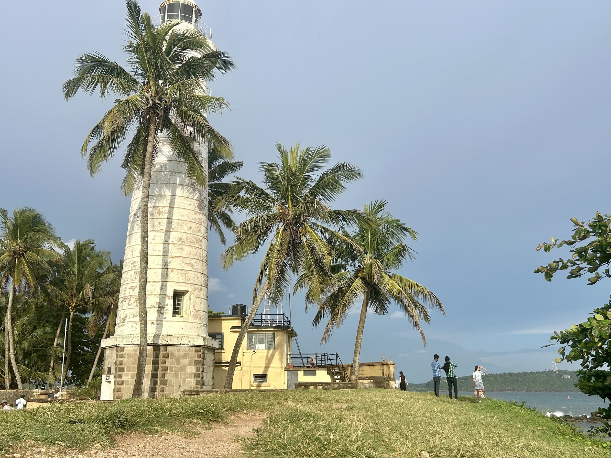 Galle lighthouse surrounded by palm trees on a grassy area near water with people walking and enjoying the view.