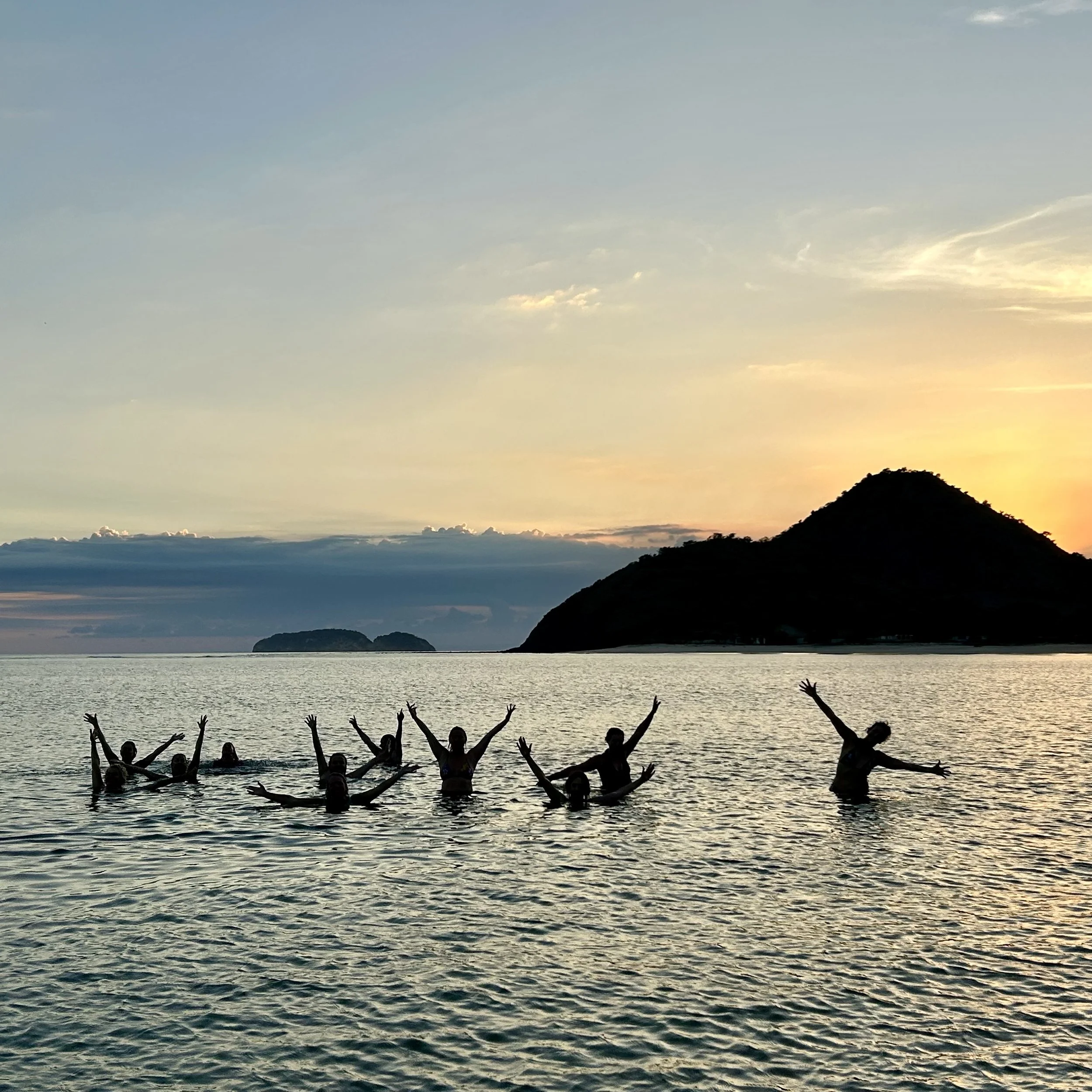 Group of people swimming and posing with arms raised in the ocean at sunset, with islands and a mountainous landscape in the background.