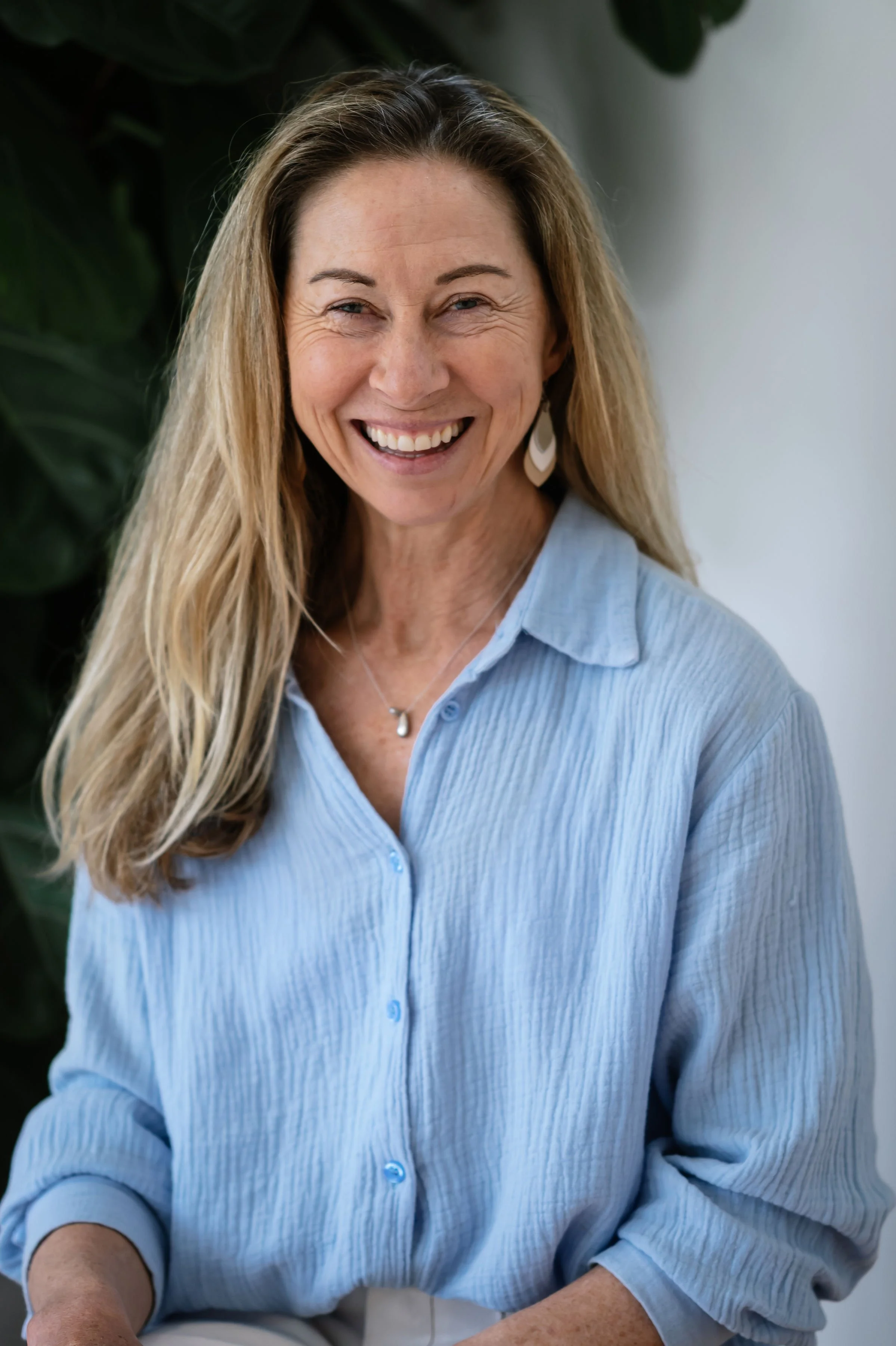 A smiling woman with long blonde hair, wearing a light blue button-up blouse and drop earrings, standing indoors with a leafy plant in the background.