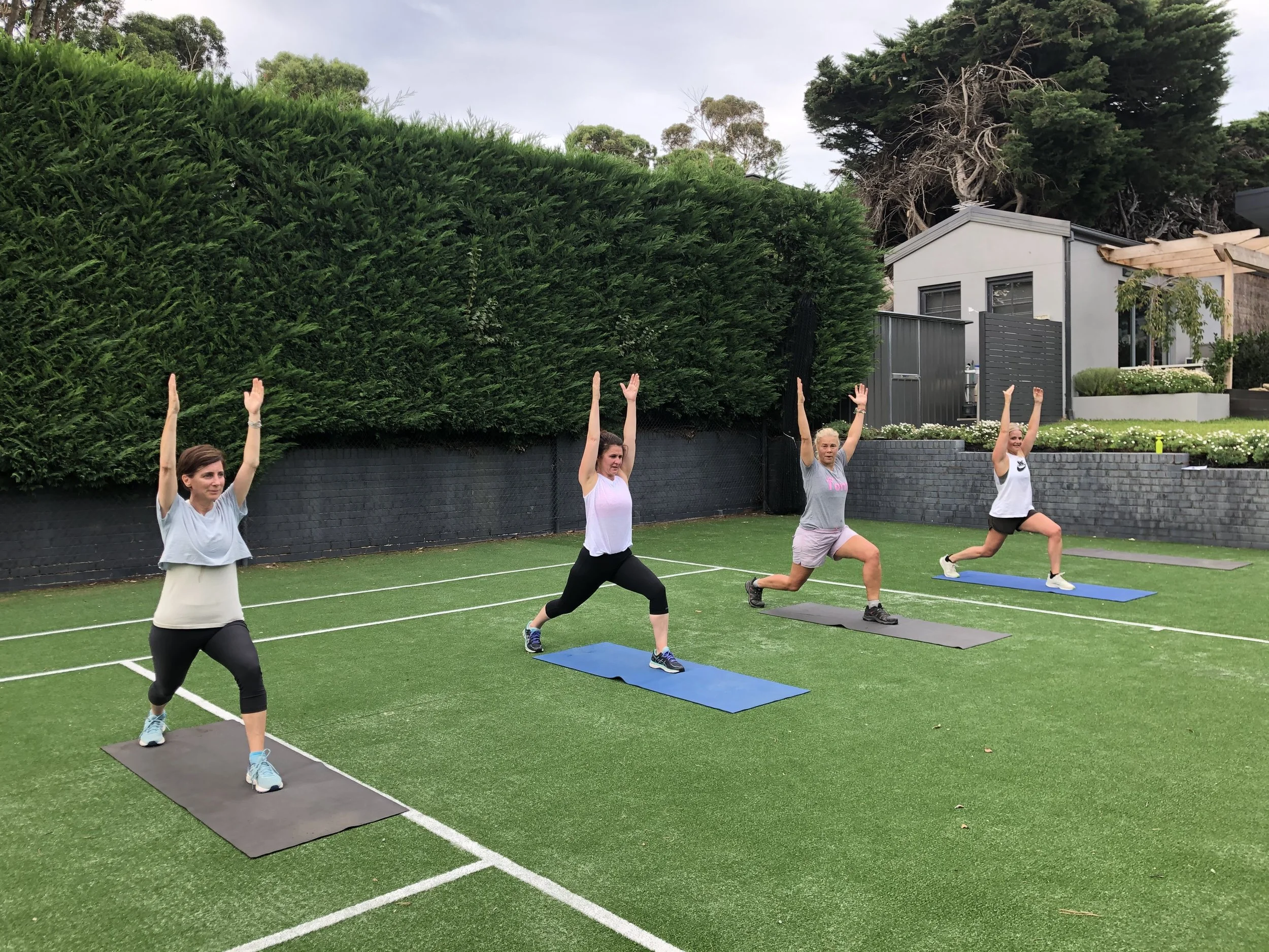 Four women exercising outdoors with Fab Over 40 Wellbeing on yoga mats, performing a yoga pose with arms raised overhead on a grassy tennis court surrounded by trees and residential buildings. Driftwood studio Mt Eliza. 