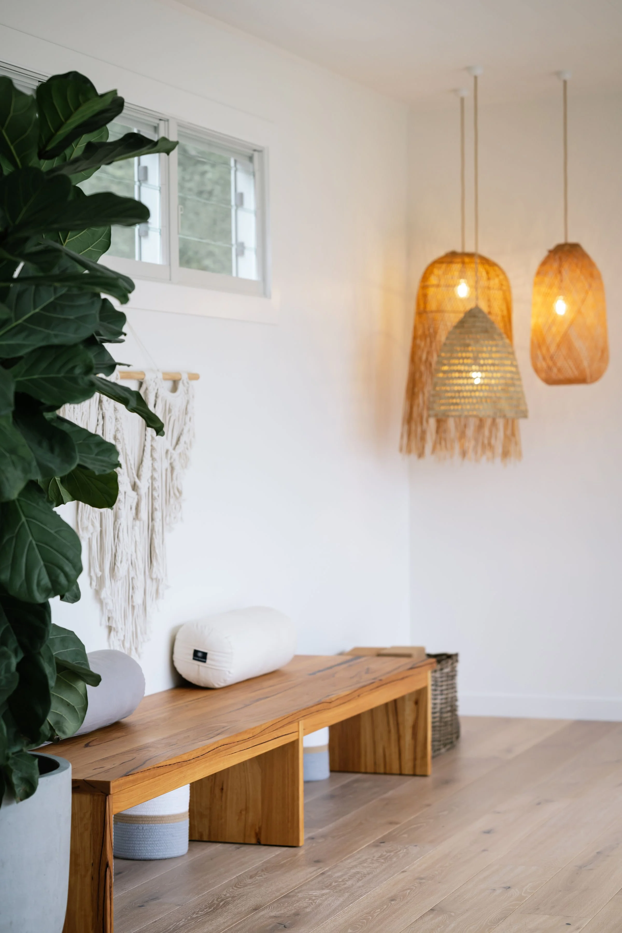 A minimalist interior with a wooden bench, large green plant, hanging woven pendant lights, a macramé wall hanging, and a window with a view of trees.