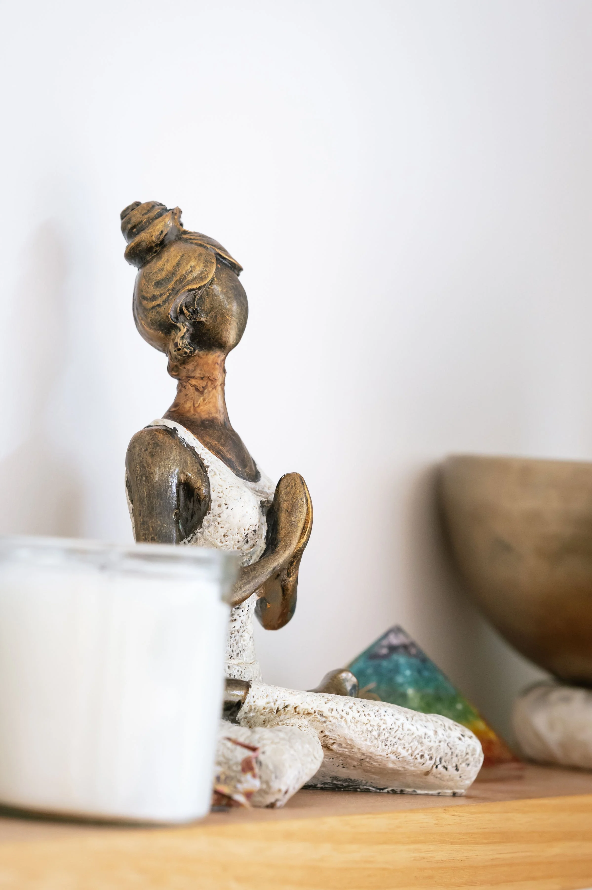 A decorative sculpture of a woman sitting cross-legged with closed eyes, holding a book, surrounded by colorful stones and a bowl, on a wooden surface. Driftwood Studio Mt Eliza. Yoga. 