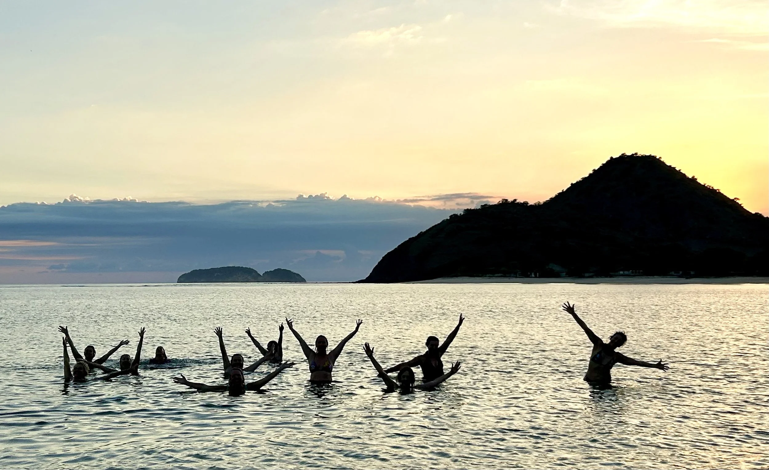 Group of people swimming and posing in the ocean at sunset with a hill and small island in the background.