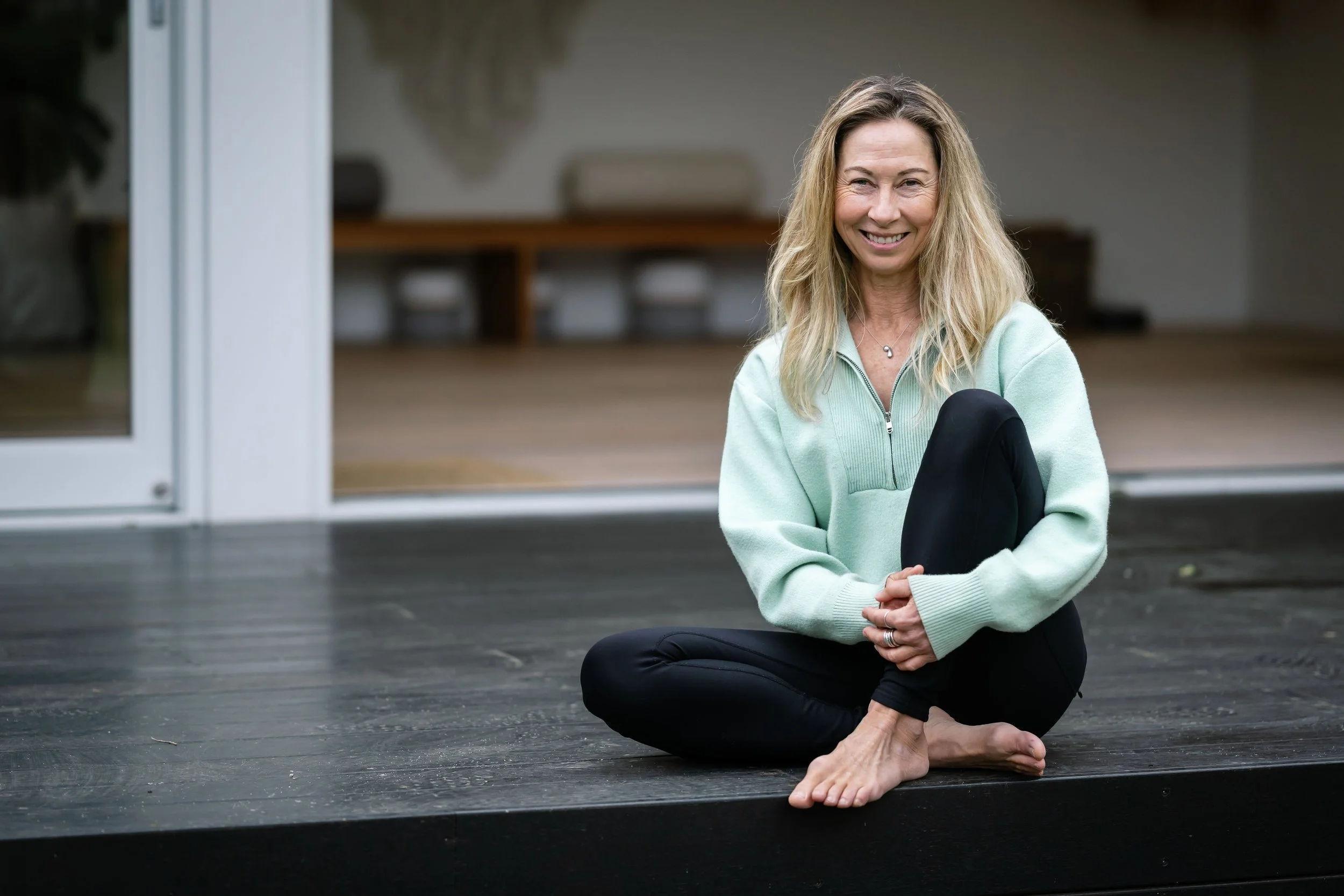 A woman sitting cross-legged on a dark wooden floor, holding a black yoga mat, smiling at the camera, with a blurred indoor background.
