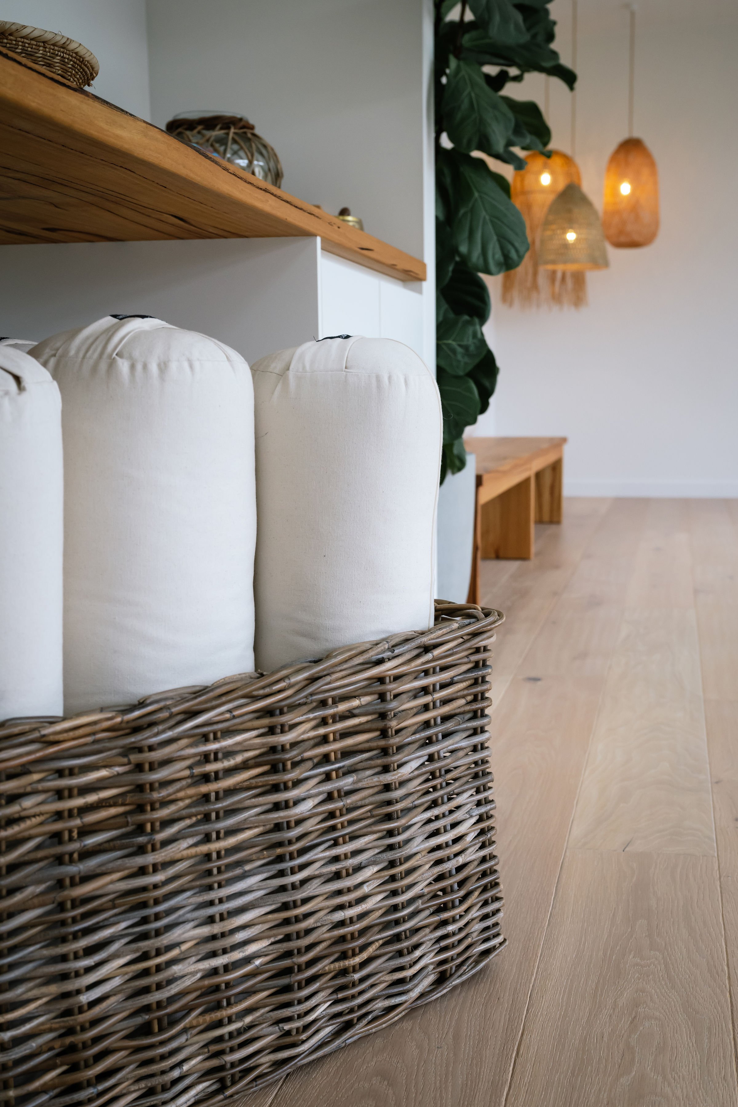 A close-up view of a wicker basket filled with rolled white cushions or pillows, placed on a light wooden floor in a modern, well-decorated interior space.