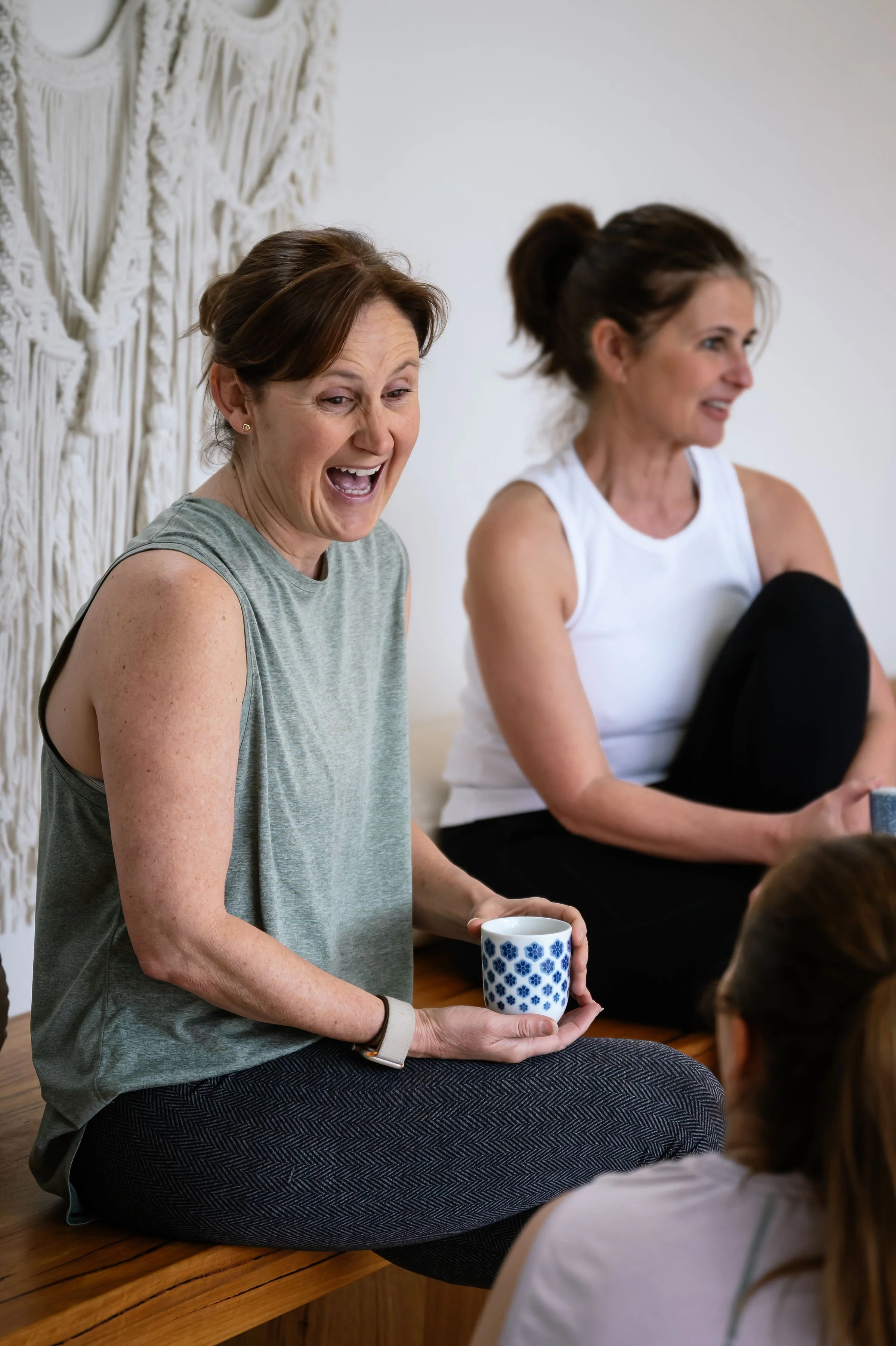 Two women sitting on a wooden bench, engaging in a lively conversation, with one holding a patterned coffee mug, in a bright room with a macramé wall hanging.