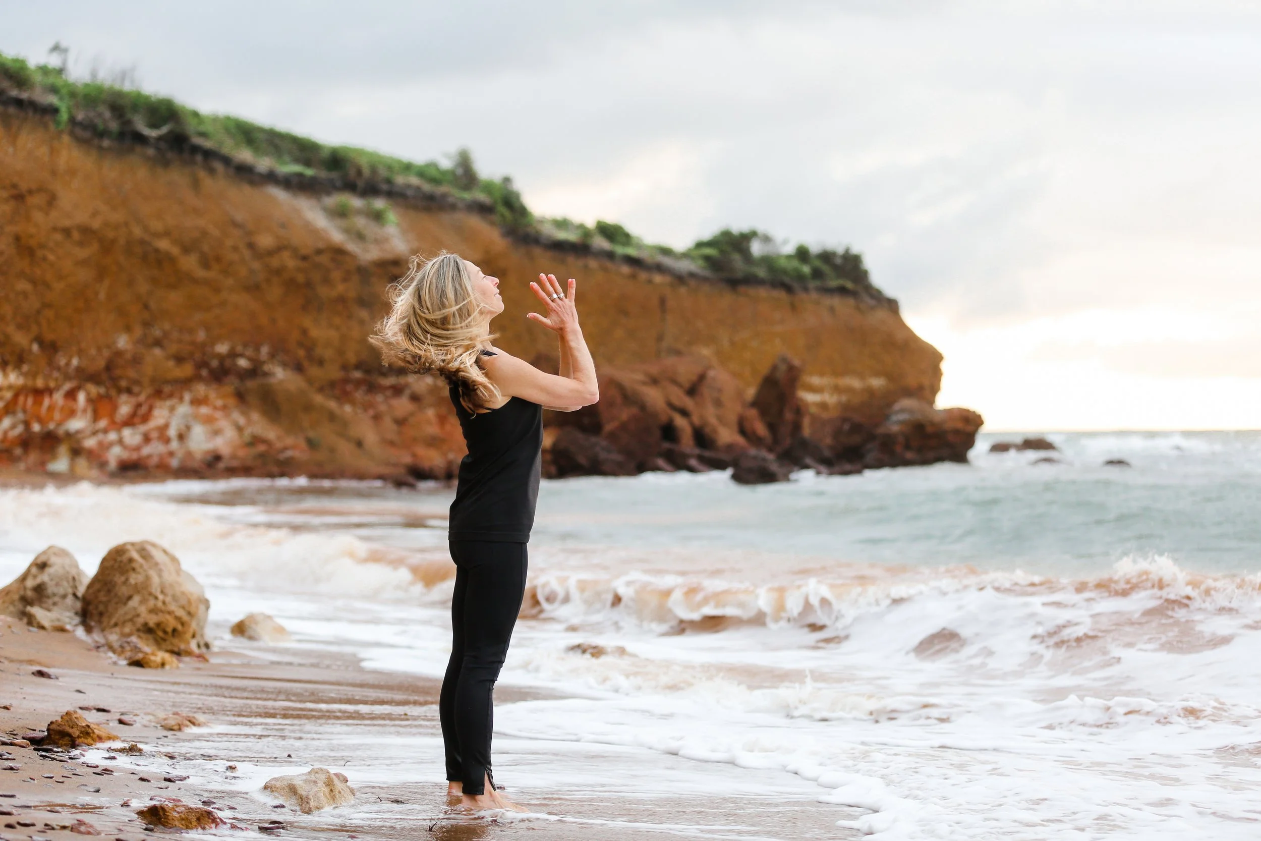 A woman practicing yoga on a beach, standing near the shoreline with waves and a rocky cliff in the background, during overcast weather.