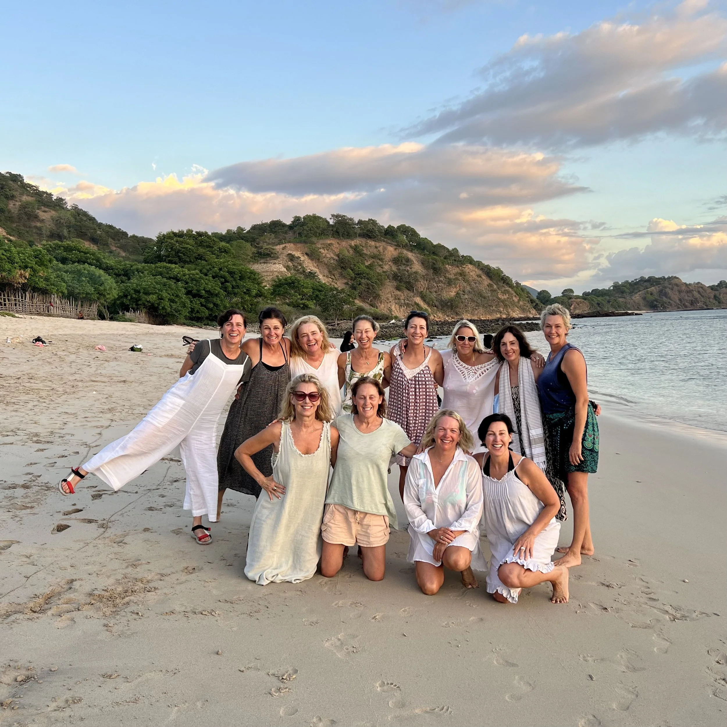 Group of women posing on a beach at sunset, with hills and sky in the background.