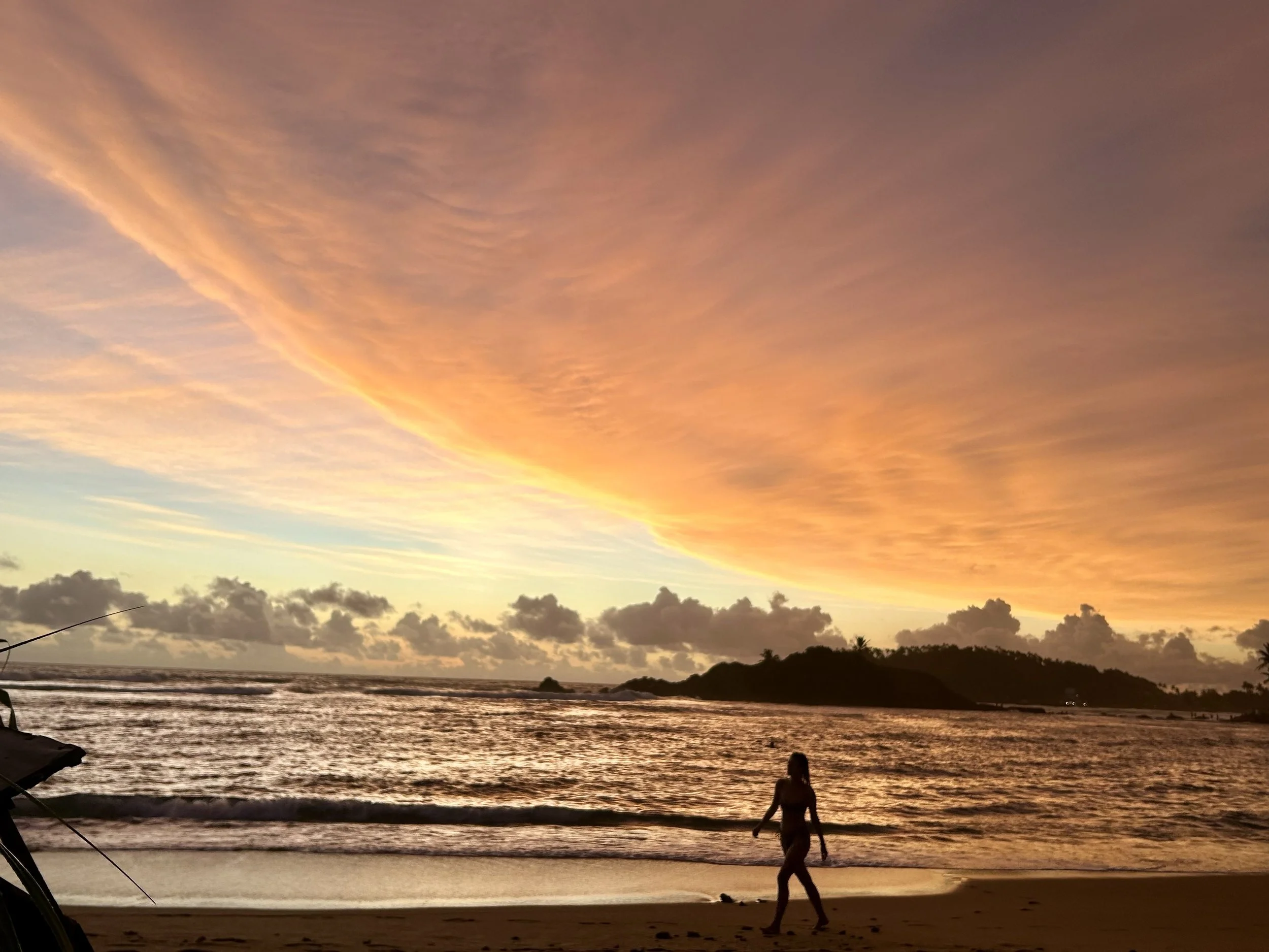 A person walking along the beach at sunset in Sri Lanka, with colorful orange and yellow clouds in the sky and an island or landmass visible in the background.