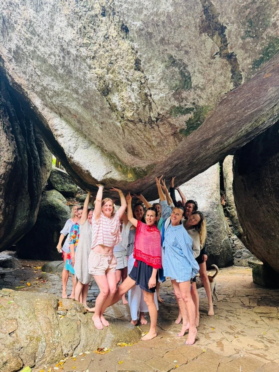 Group of people standing under a large, overhanging rock formation, smiling and raising their hands, with some interacting with a dog nearby.
