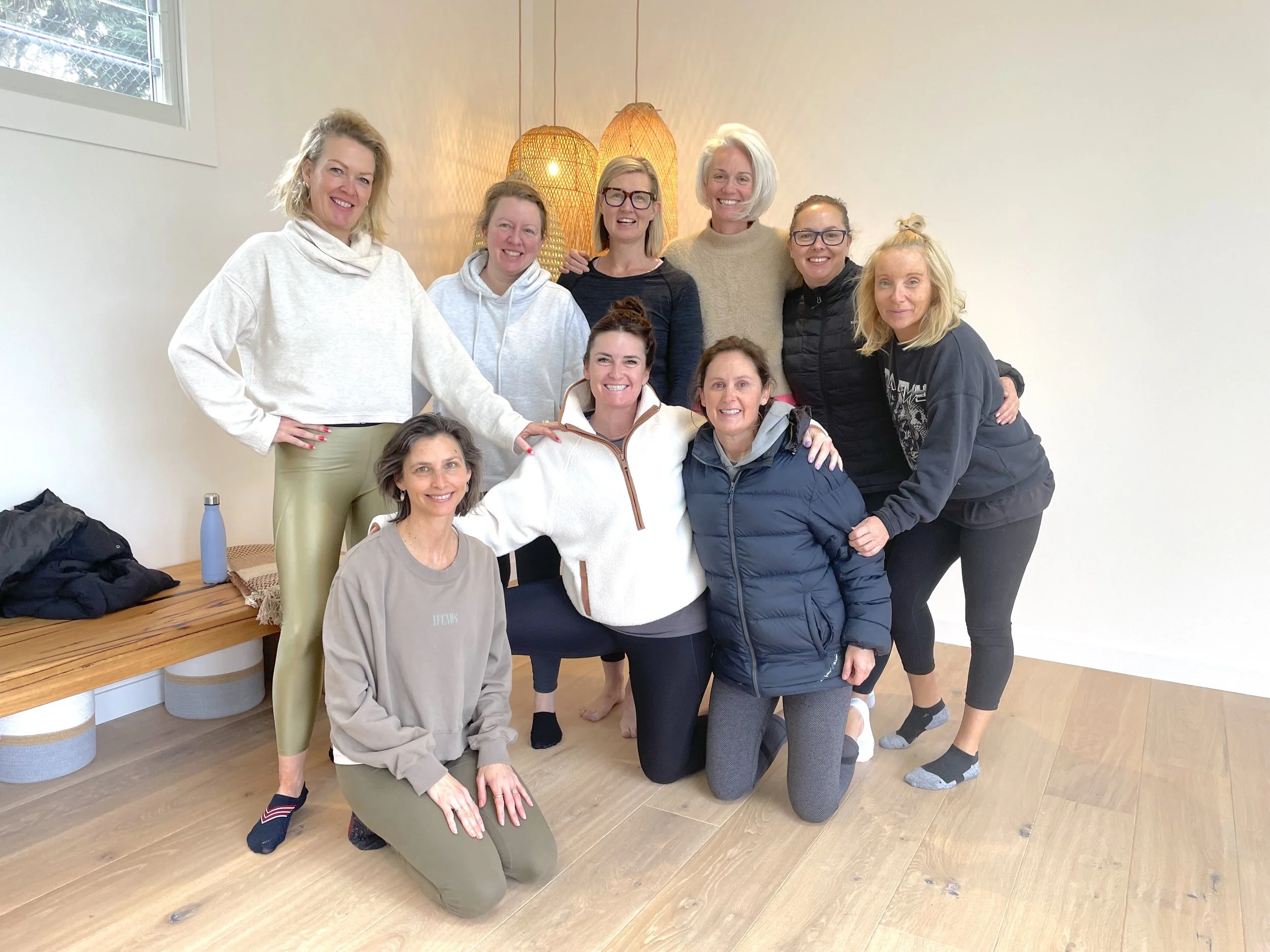 Group of ten women gathered indoors, some standing and some kneeling, smiling for the camera. They are in a room with wooden flooring, a white wall, a window, and a wooden bench with items on it. Four women are wearing casual workout or lounge clothe