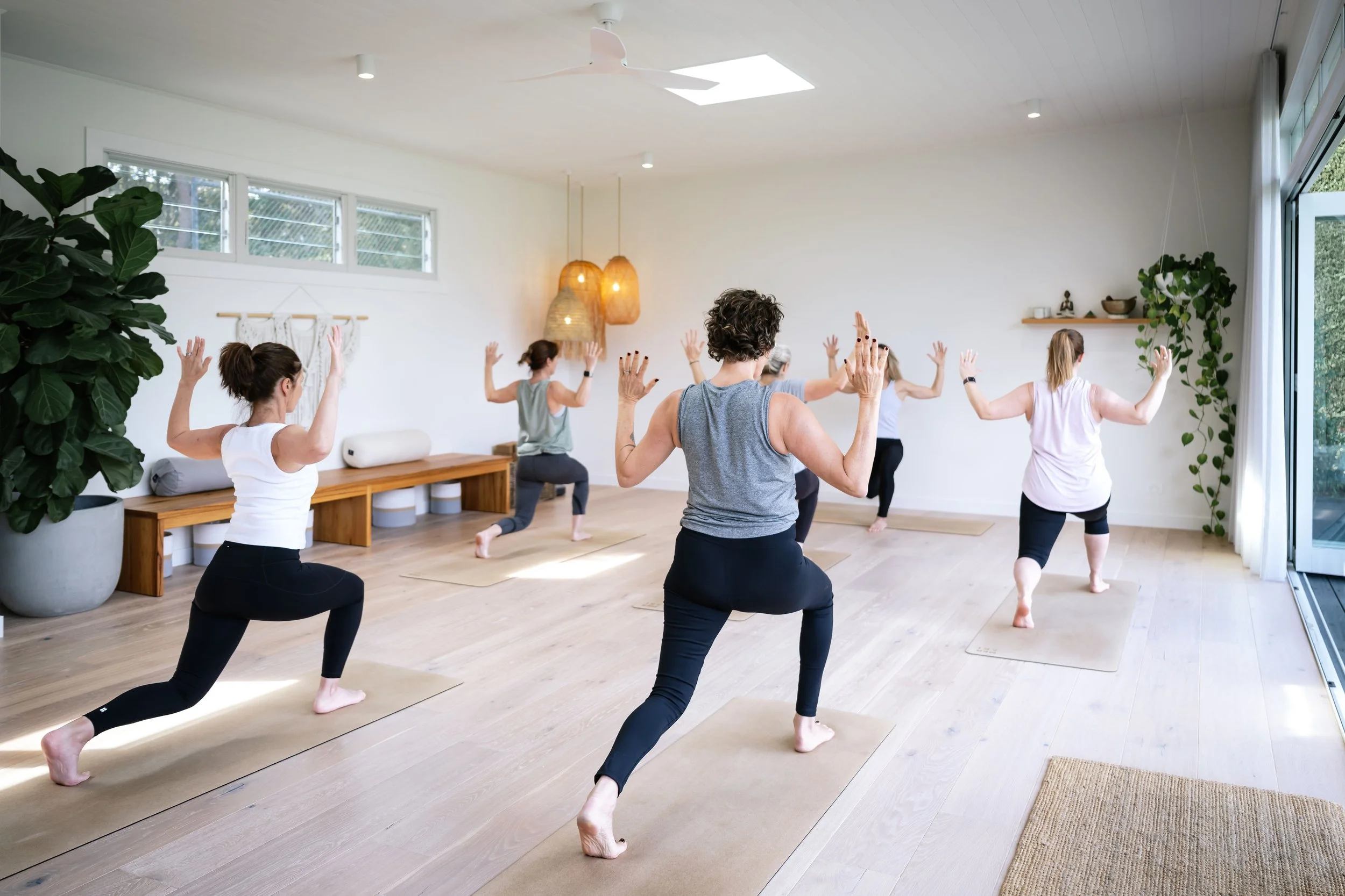Group of women participating in a yoga or meditation class in a bright, airy room with large windows, plants, and minimal decor.