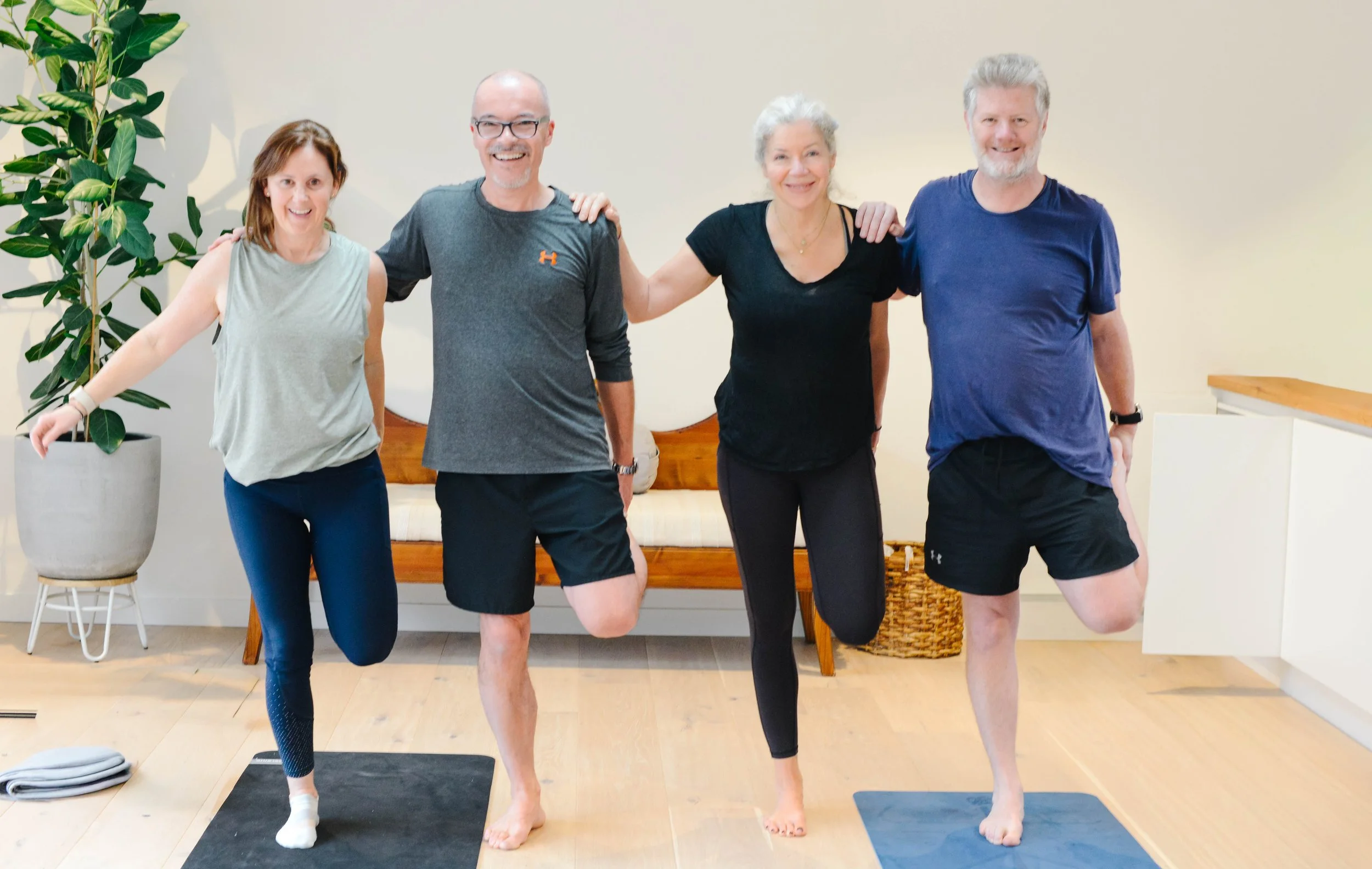 Four people practicing yoga indoors, standing on yoga mats, holding one leg with their hands on their hips and smiling.