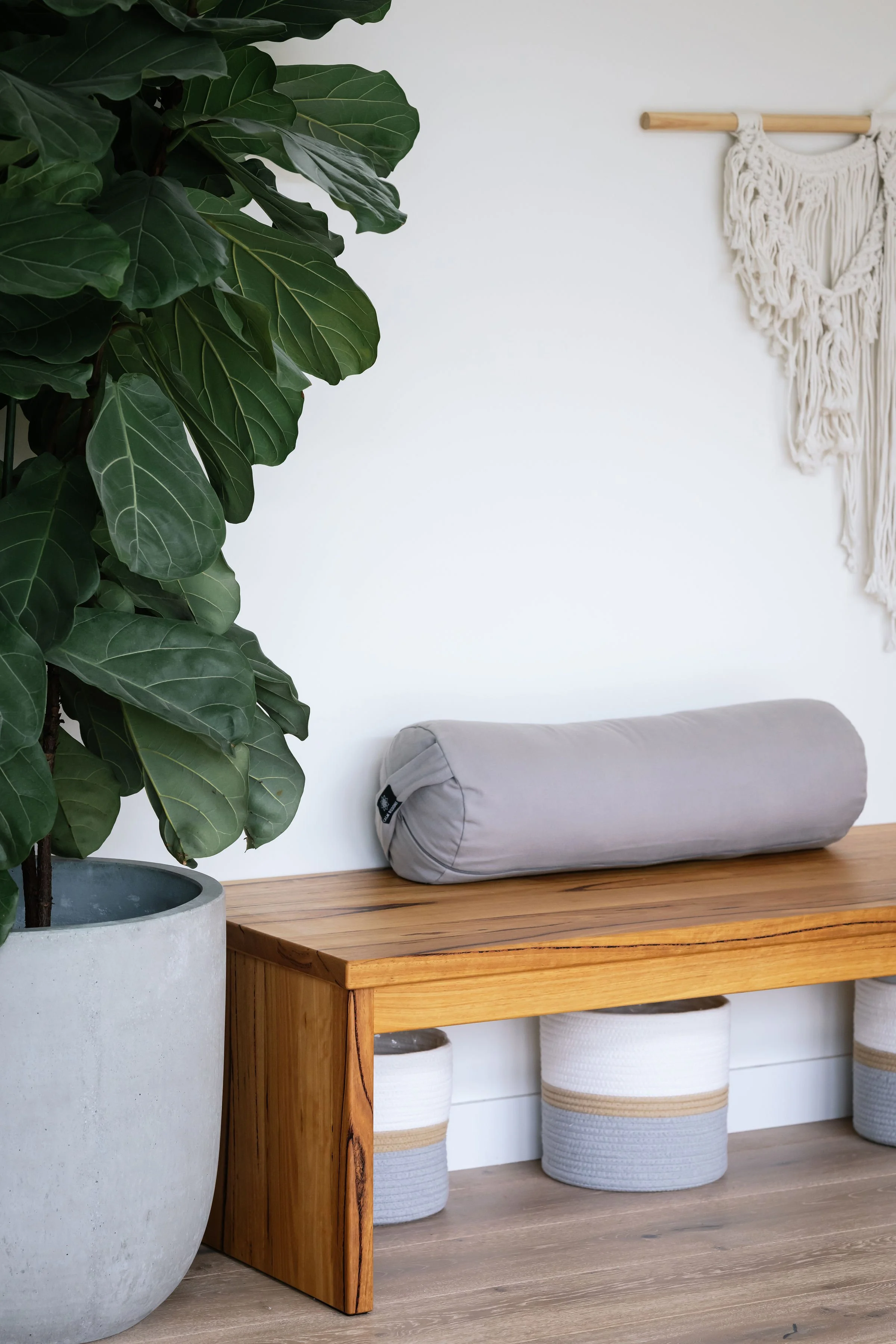 A large potted Fiddle Leaf Fig plant in Driftwood Studio Mt Eliza. There is a wooden bench with a gray bolster pillow, and three woven baskets underneath, with a white wall and a macramé wall hanging in the background.