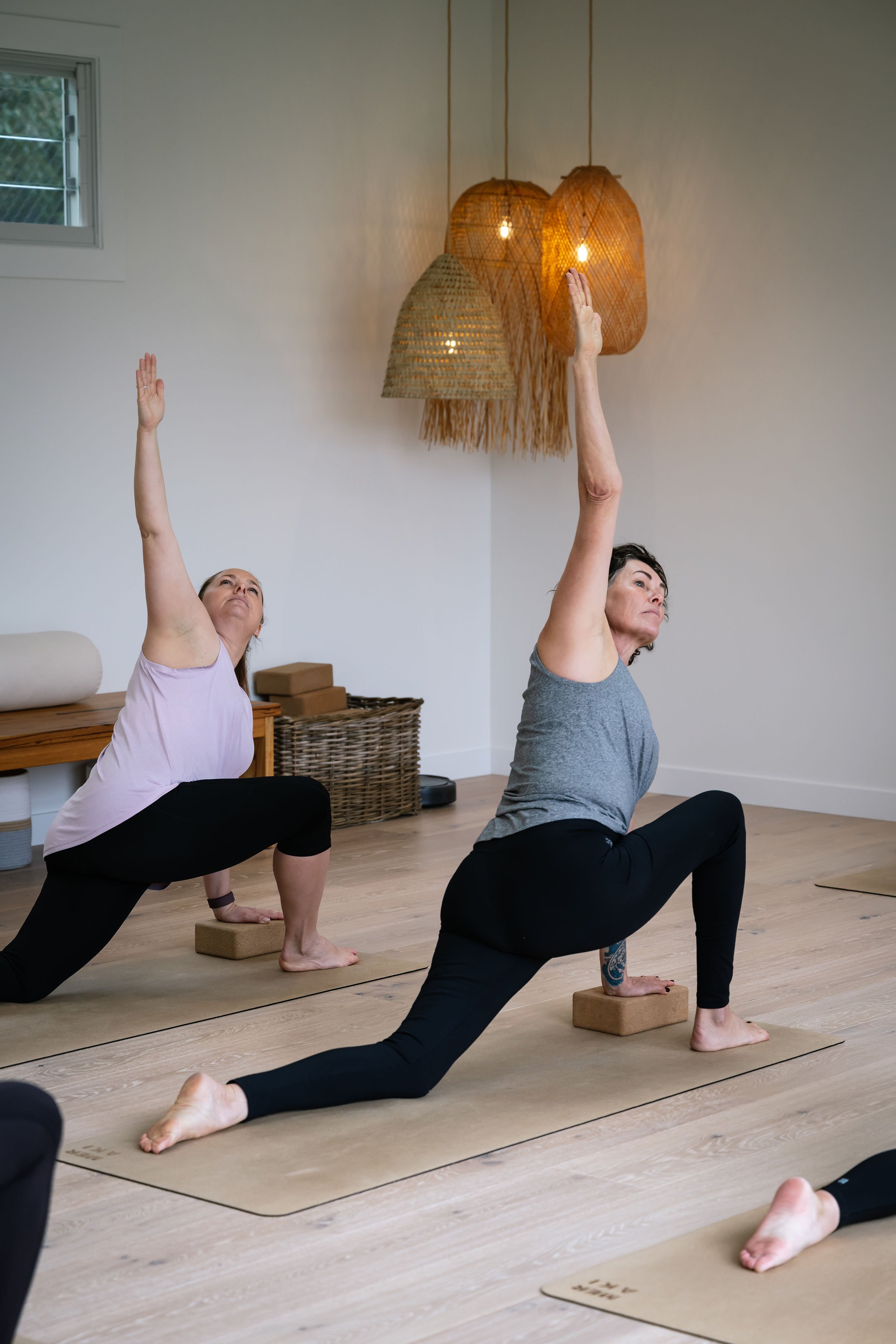 Two women practicing yoga in a studio, both in lunge position with one hand on the ground and the other reaching upward, using yoga blocks for support, with woven pendant lights hanging from the ceiling.