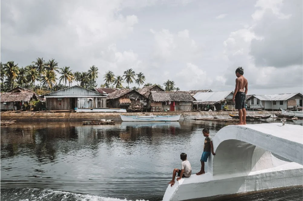 A young boy stands on a white boat docked on a river, with two children sitting on its edge, a shirtless man standing behind them, and traditional houses with thatched roofs and palm trees in the background.