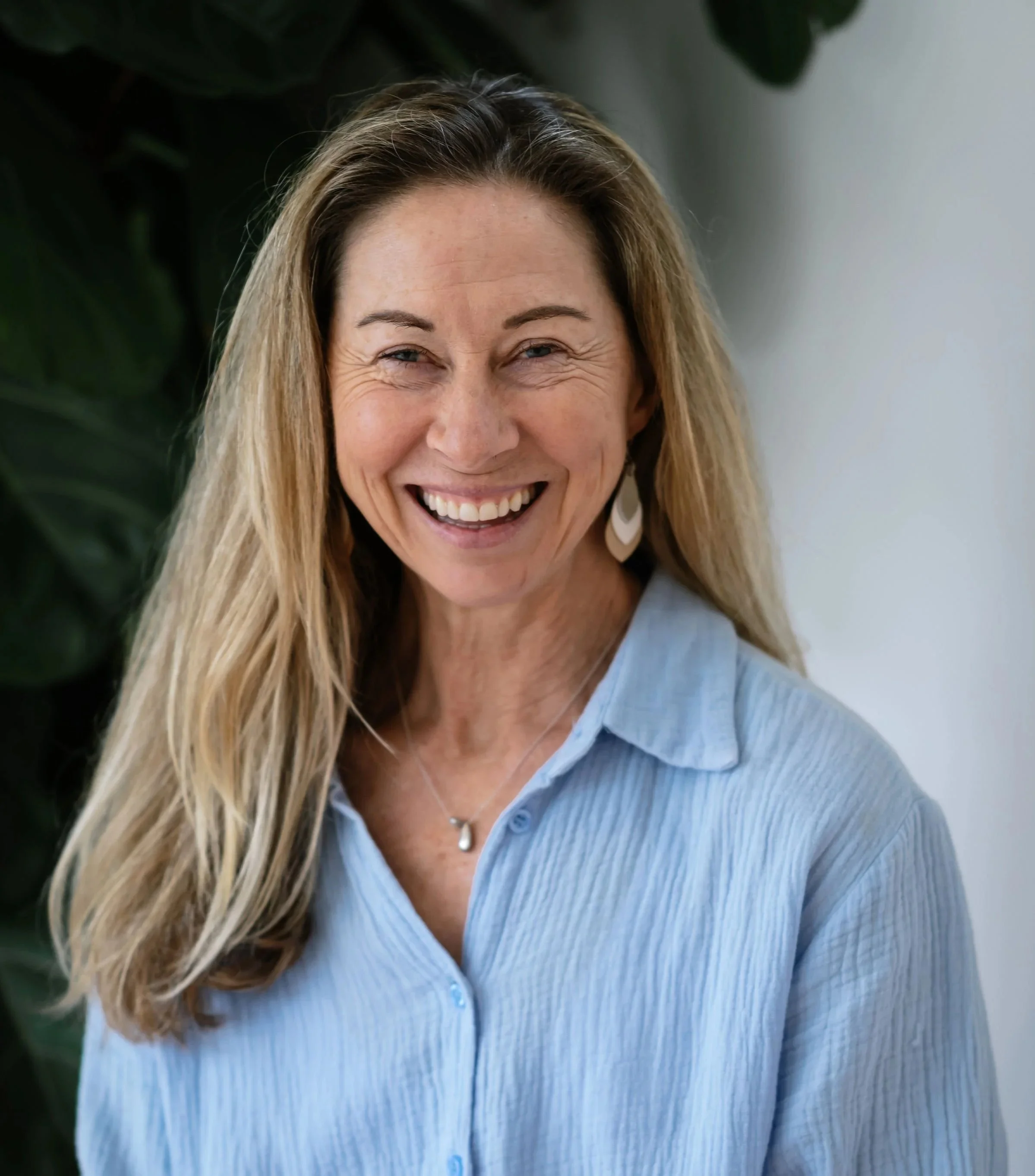 A smiling woman with long blonde hair wearing a light blue shirt standing indoors with a leafy plant in the background.