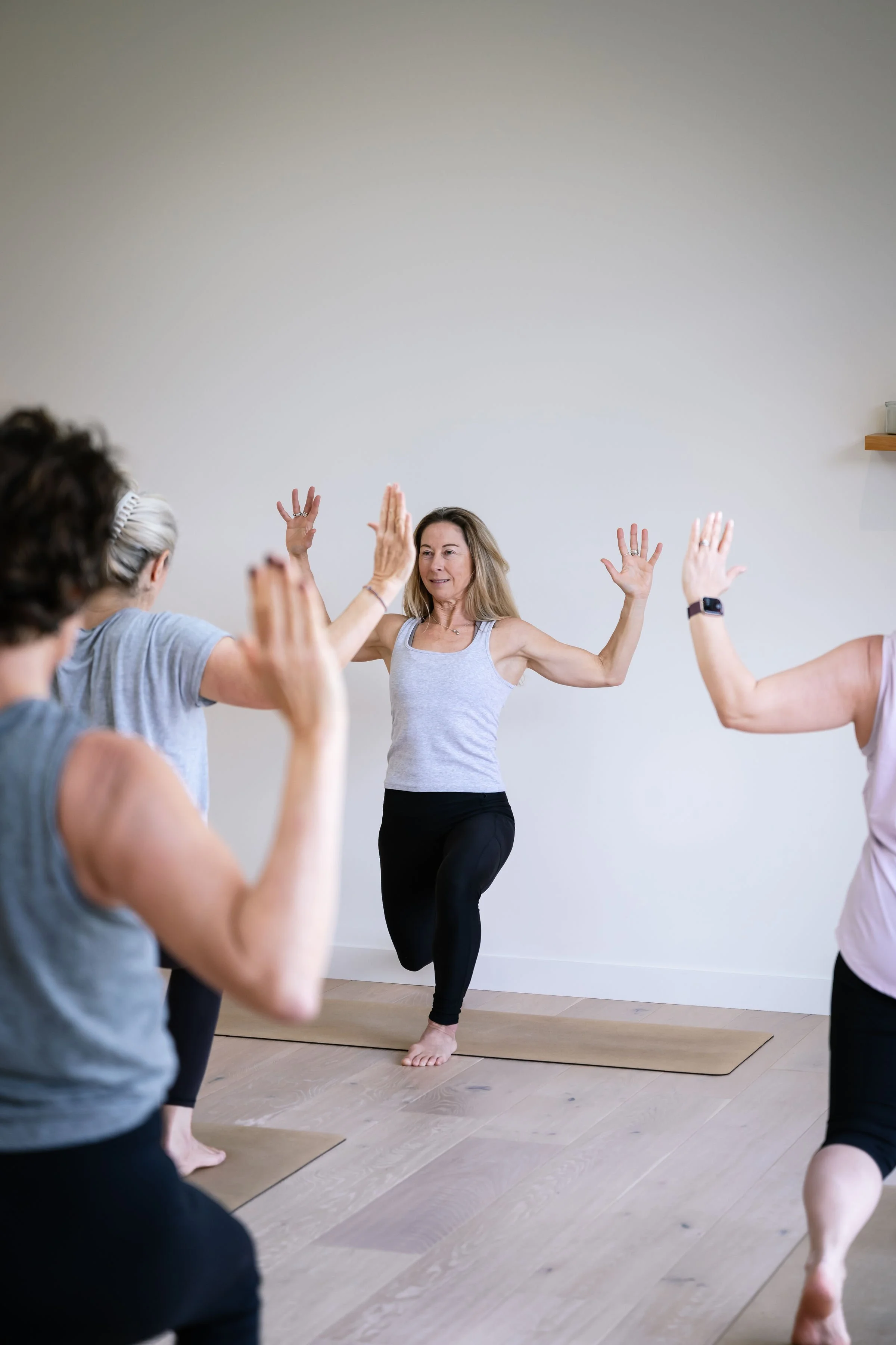 Women participating in a yoga class, with a woman in the center demonstrating a pose with arms raised and one foot lifted, on yoga mats in a room with a plain white wall.