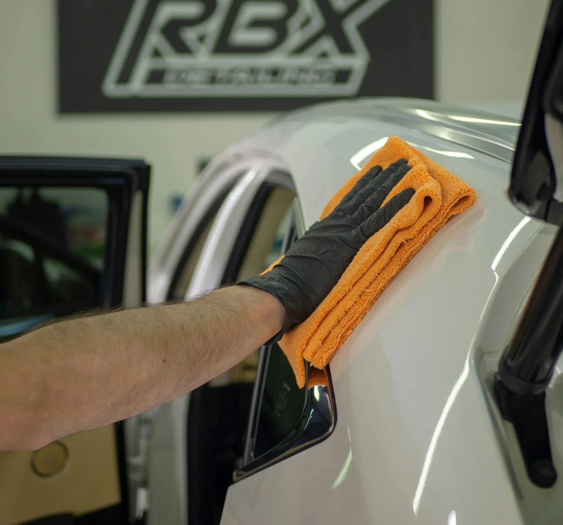 A person cleaning a white car's door handle area with a plush orange microfiber cloth, wearing a black glove on their hand.