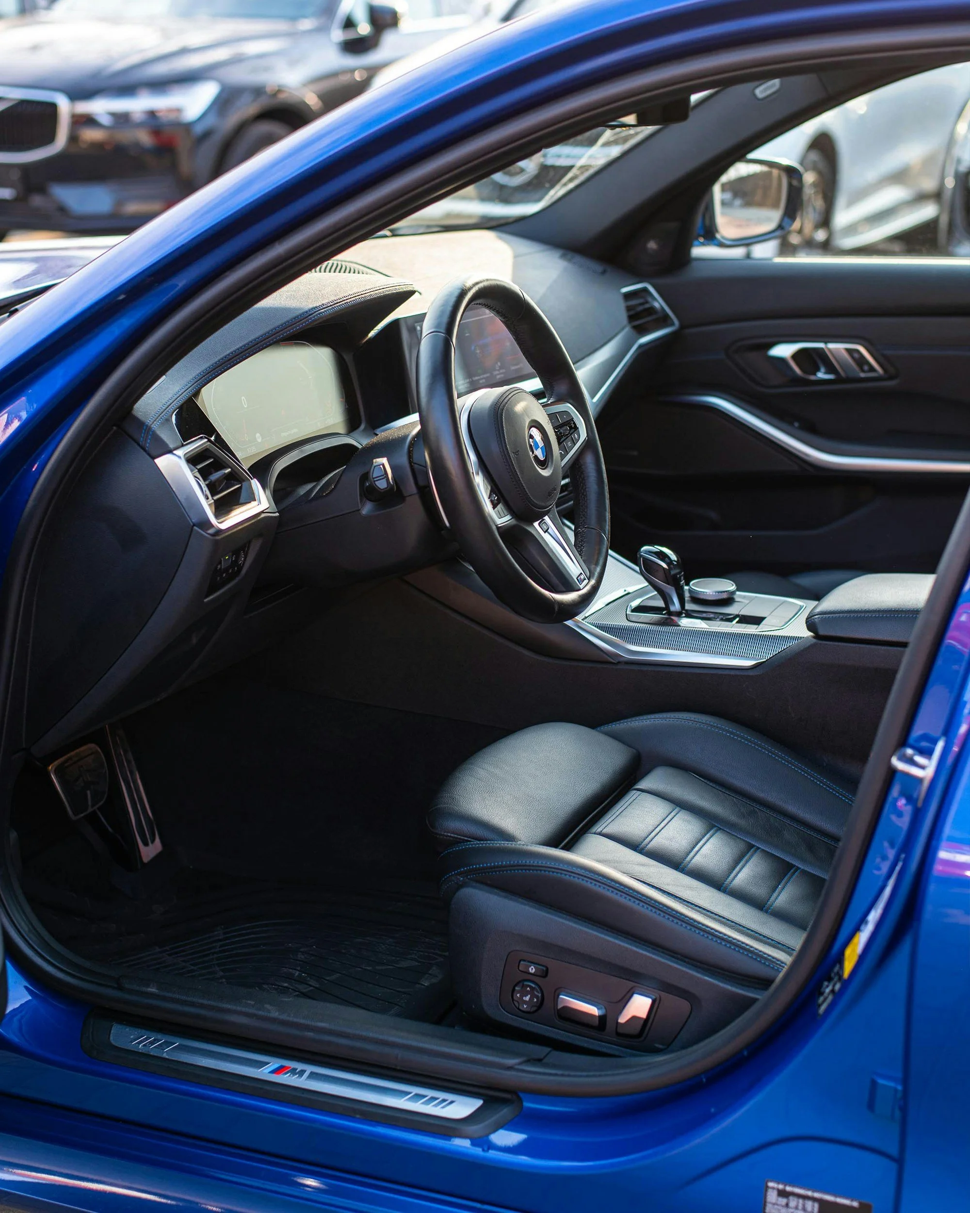 Interior of a blue BMW car showcasing the steering wheel, dashboard, gear shifter, and driver's seat with controls, parked in a lot with other cars visible outside.