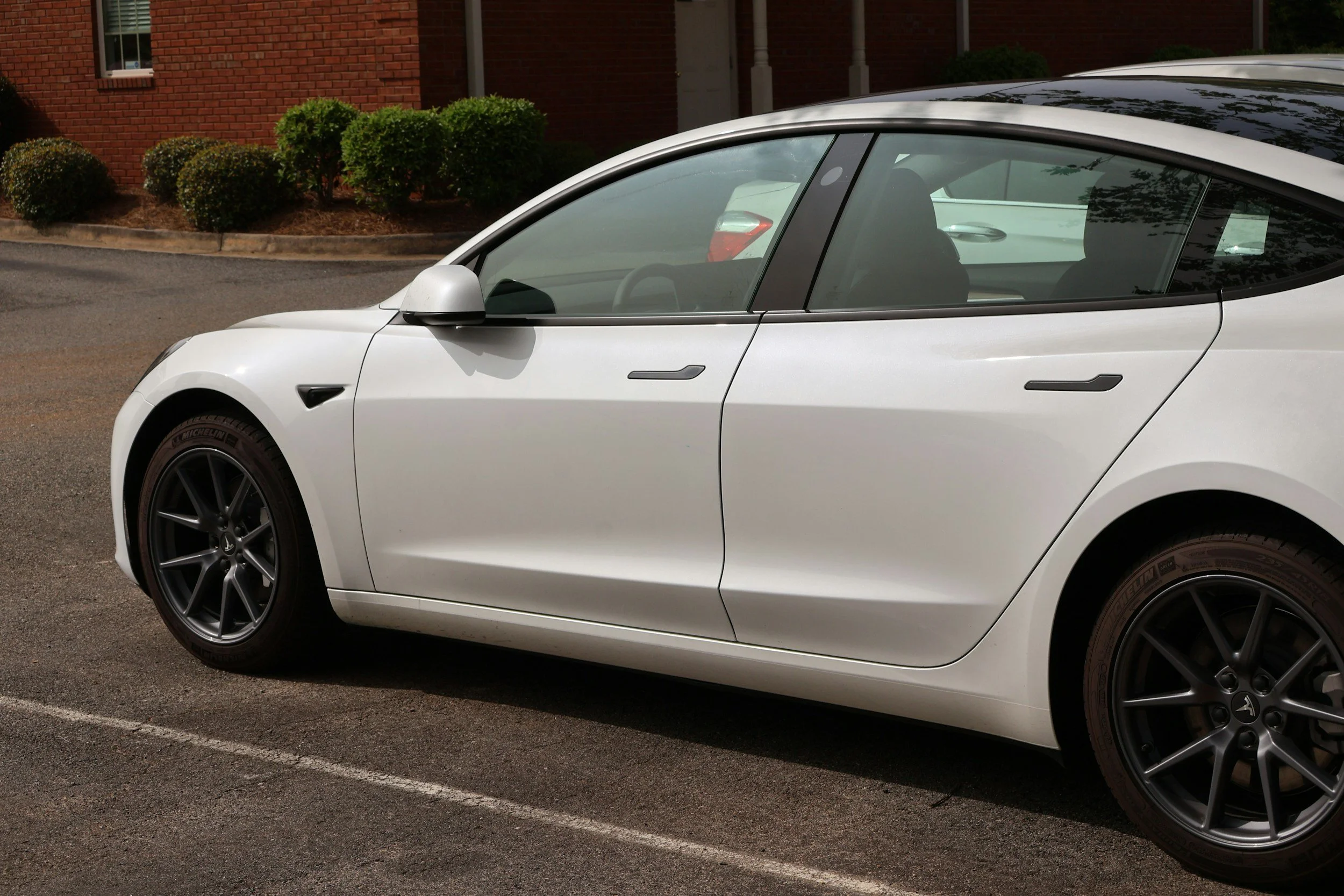 A clean white Tesla Model 3 parked in a parking lot near a brick building with bushes.