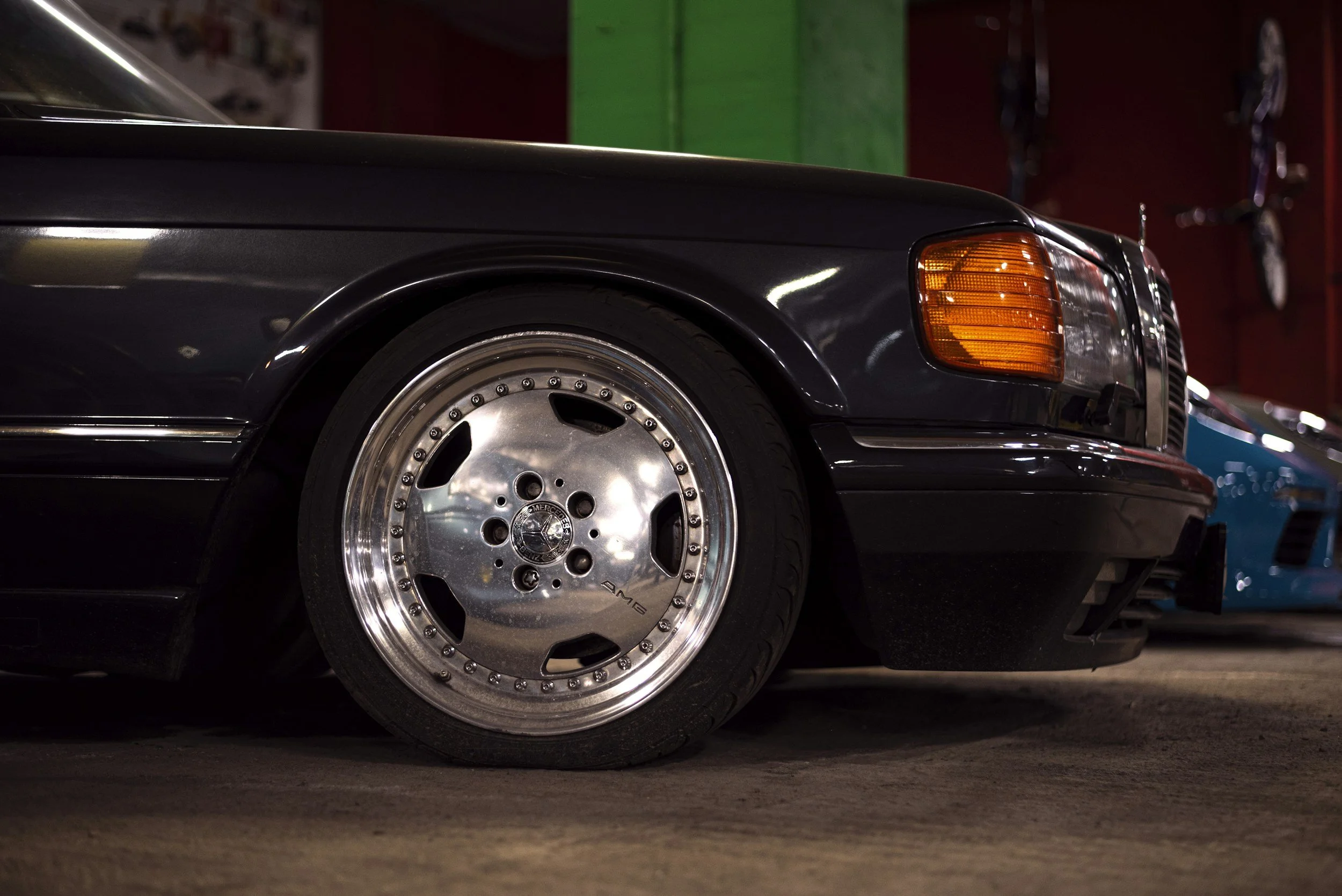 Close-up of a black vintage Mercedes-Benz car with chrome AMG wheels inside a garage, with another blue sports car visible in the background.