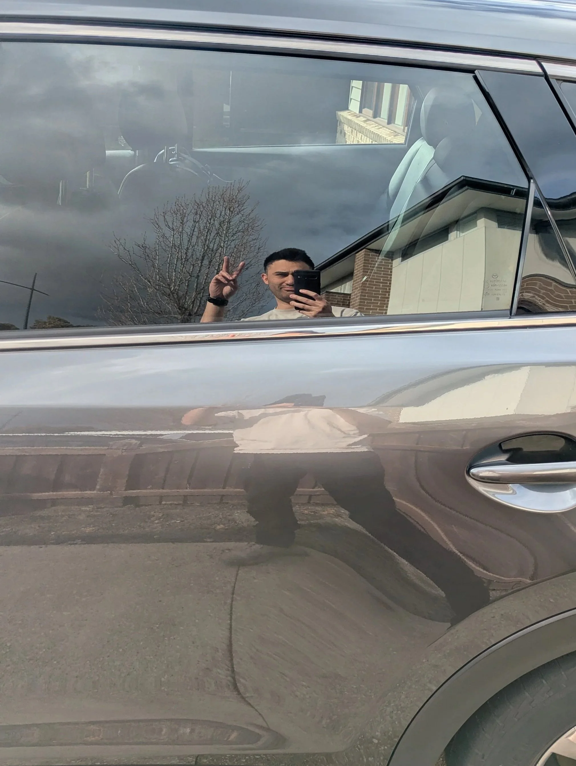 Reflection of a man taking a selfie in a shiny car window, making a peace sign with his hand, with cloudy sky and building in the background.