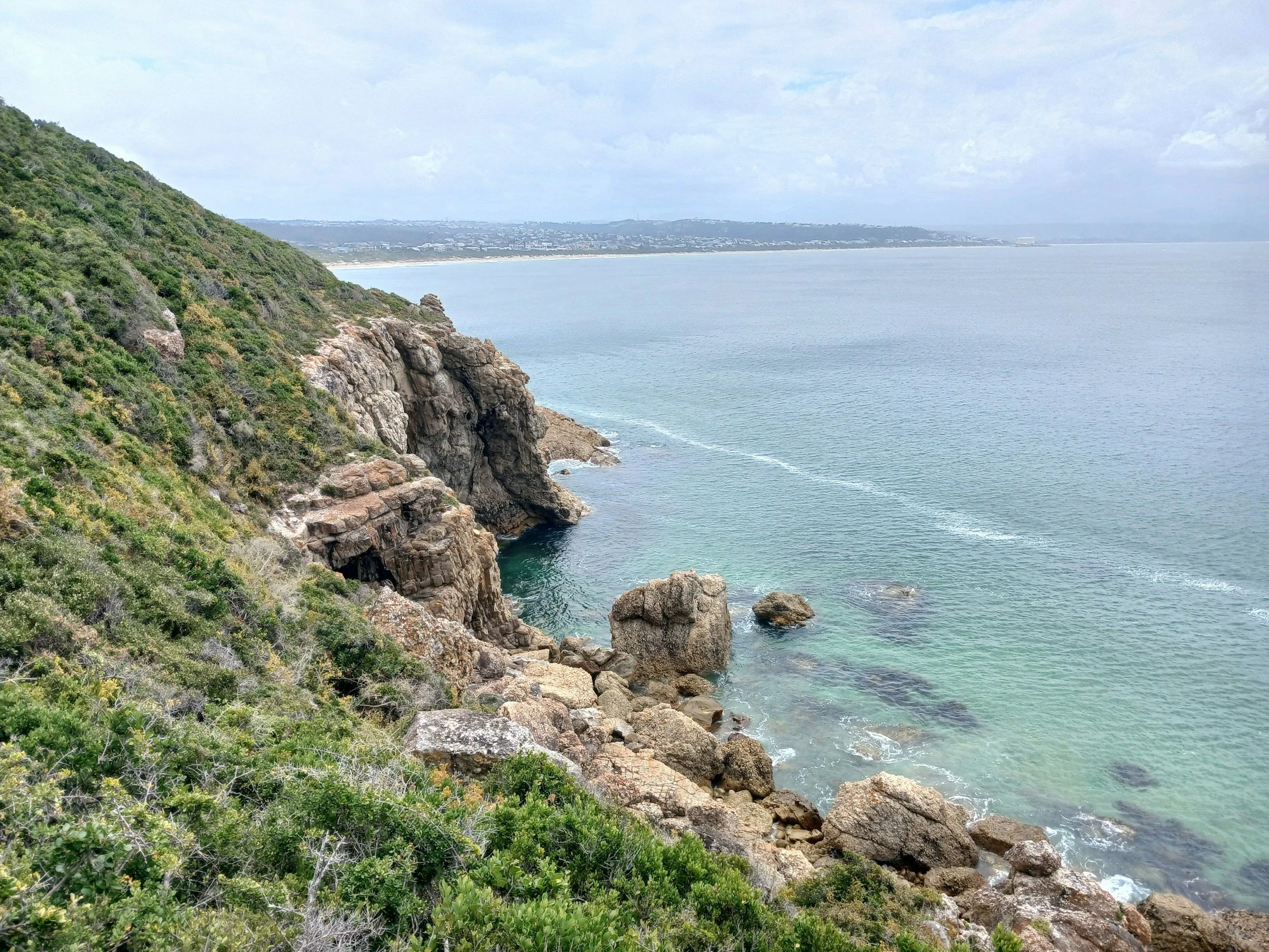 A scenic coastal landscape with rocky cliffs and lush green vegetation on the left, leading down to clear, turquoise water with some rocks visible beneath the surface. The ocean extends towards a distant shoreline under a partly cloudy sky.