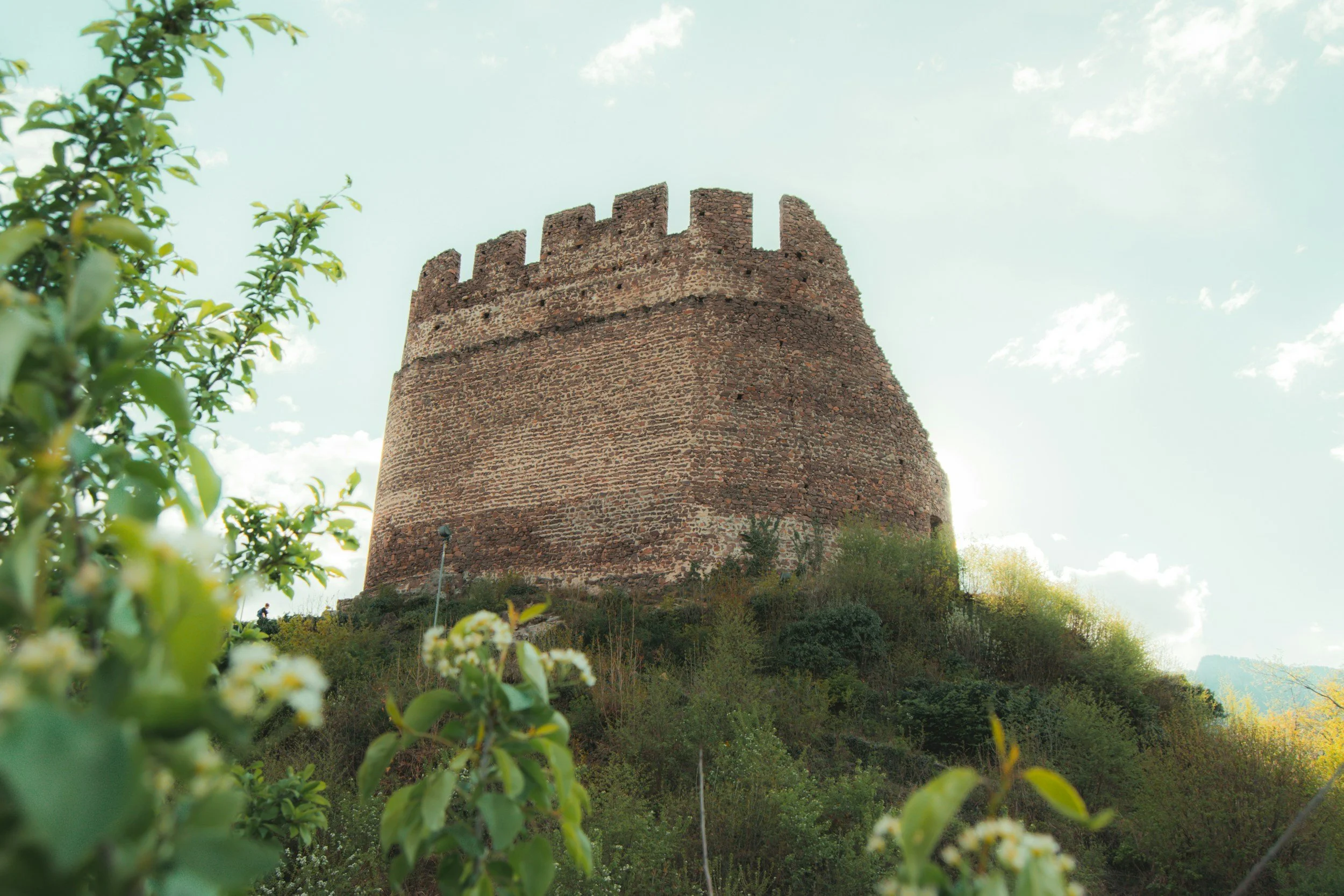 A tall, ancient brick tower on a hill surrounded by green vegetation, with a partly cloudy sky in the background.