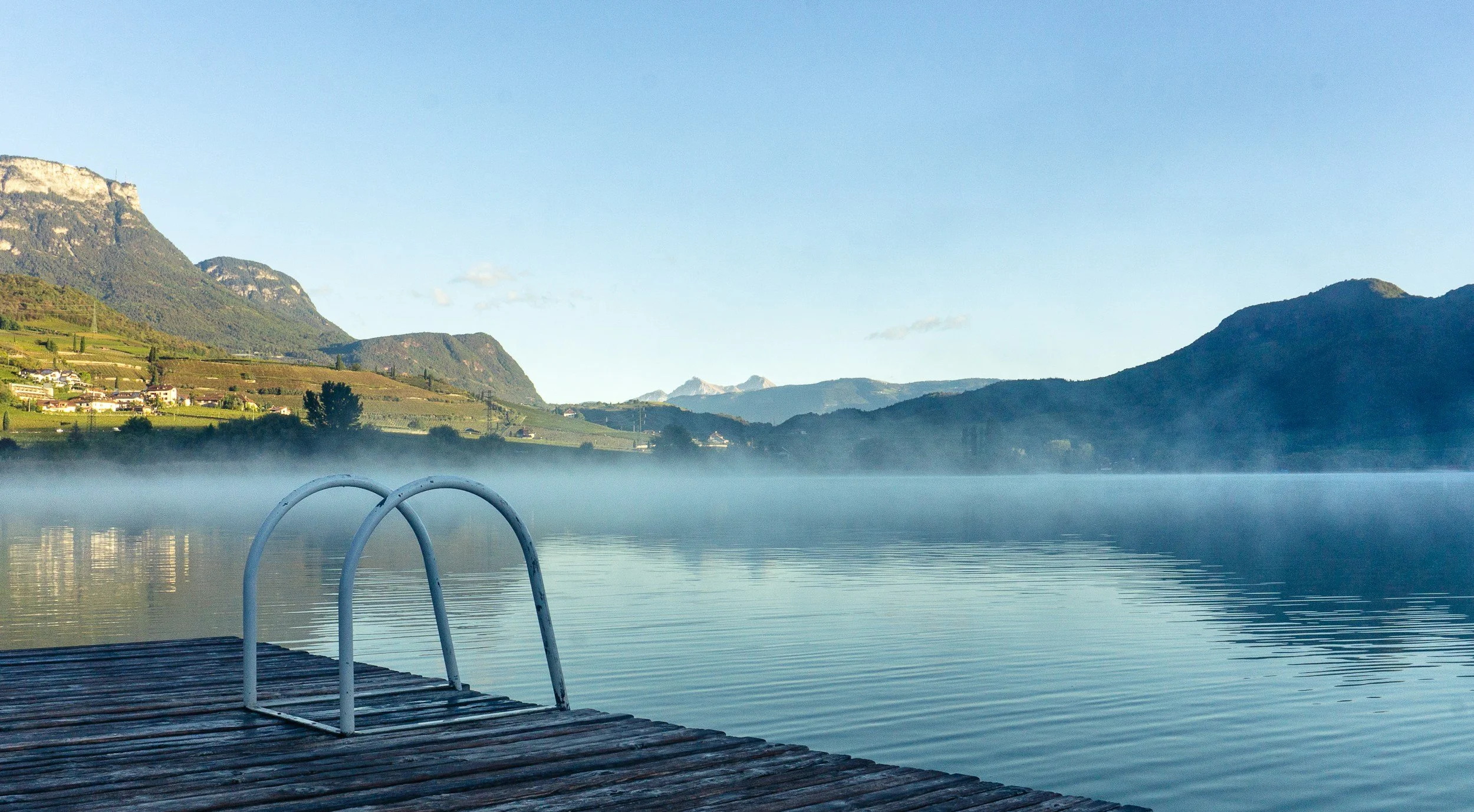 A wooden dock with a metal ladder extending into a calm lake, surrounded by mountains under a clear blue sky with some mist over the water