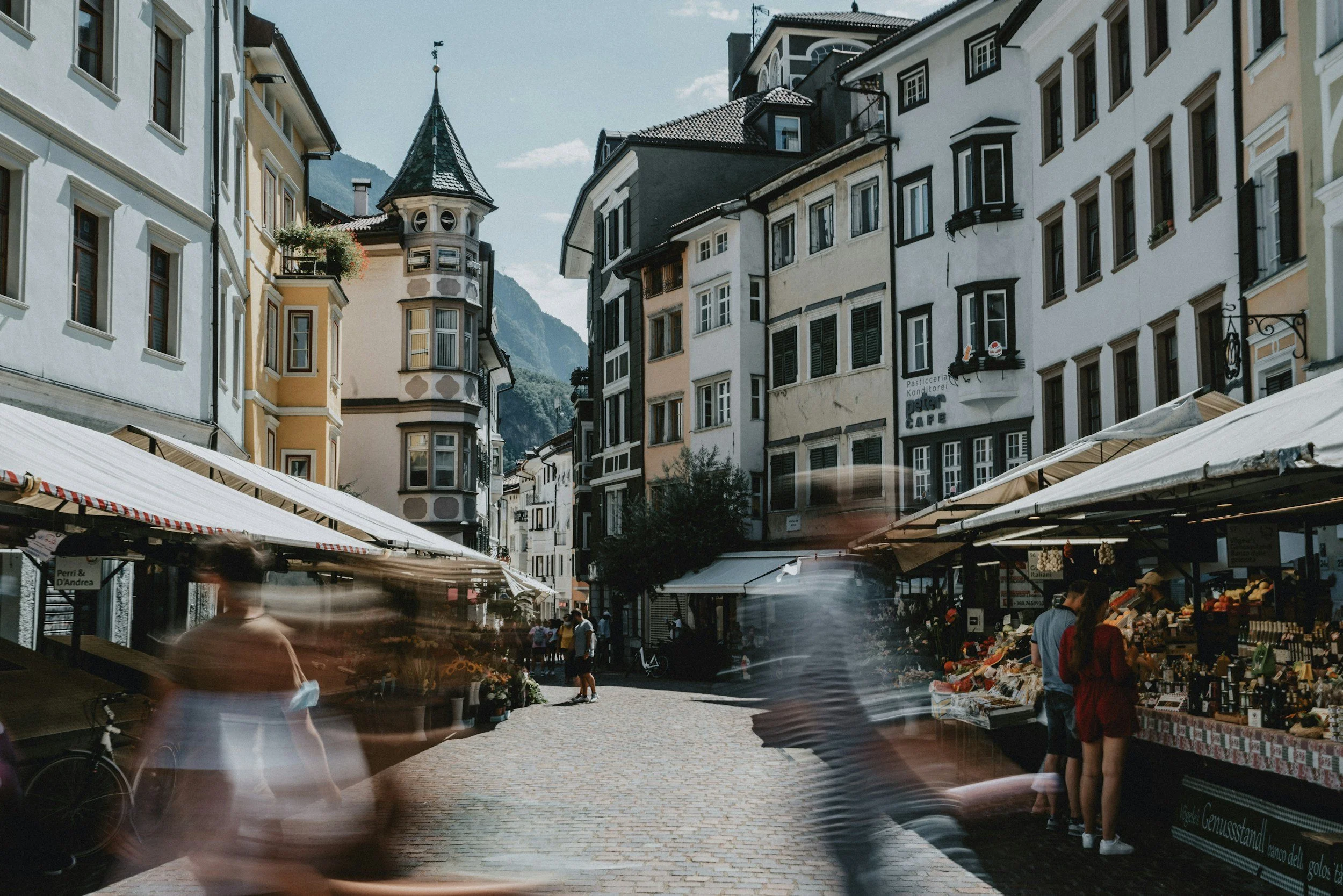 A street in a European town with market stalls, people walking, and colorful old buildings with mountains in the background.