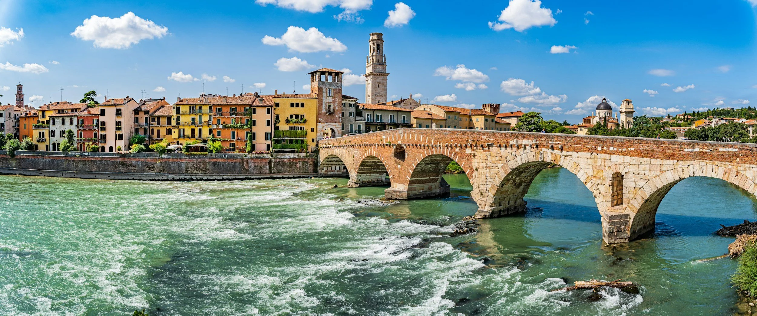 A scenic view of a stone arched bridge crossing a river with gentle rapids, in front of colorful Italian buildings under a bright blue sky with a few scattered clouds.