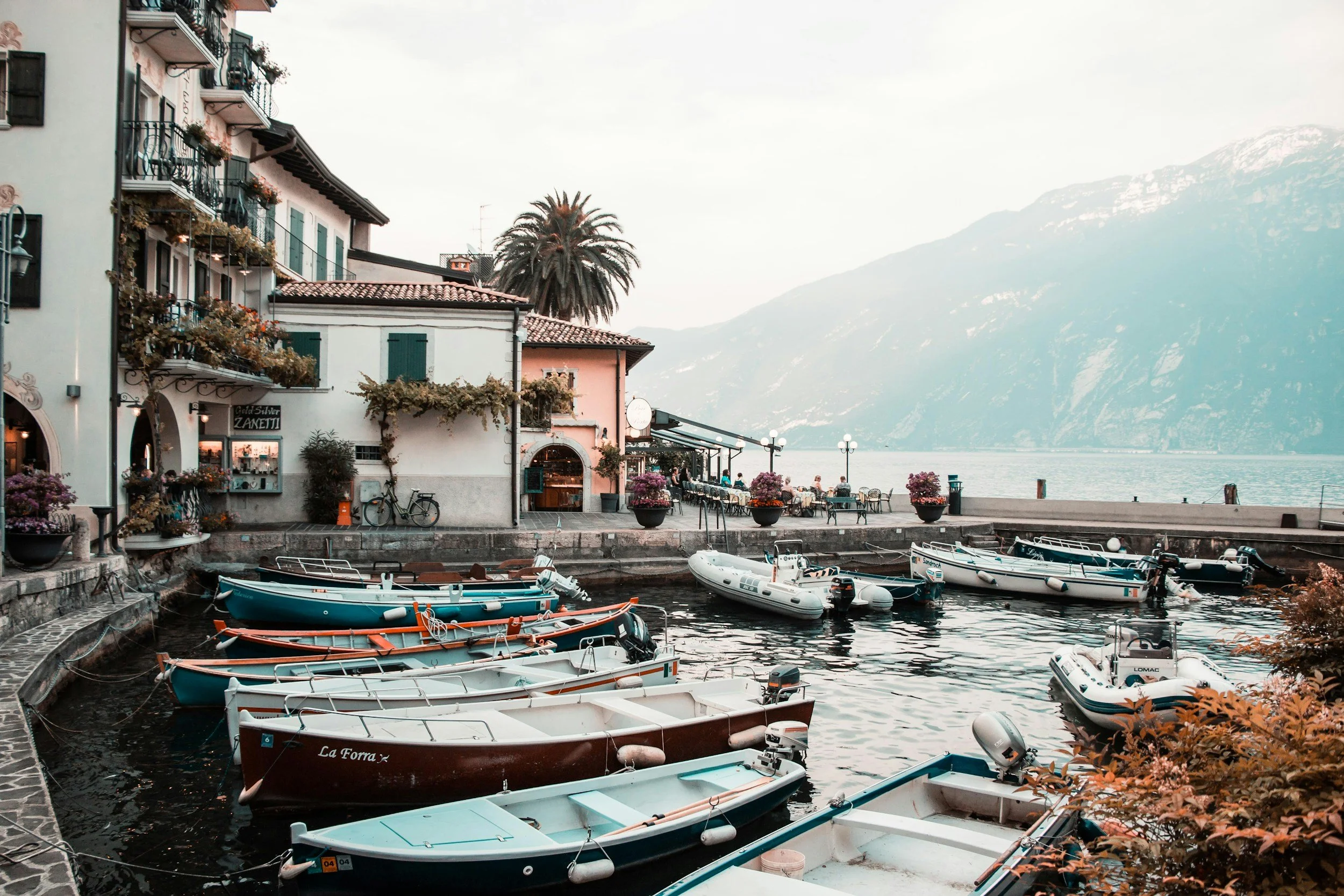 A picturesque harbor with several boats docked along a stone pier, colorful buildings with balconies decorated with plants on the left, and a mountainous landscape with a lake in the background.