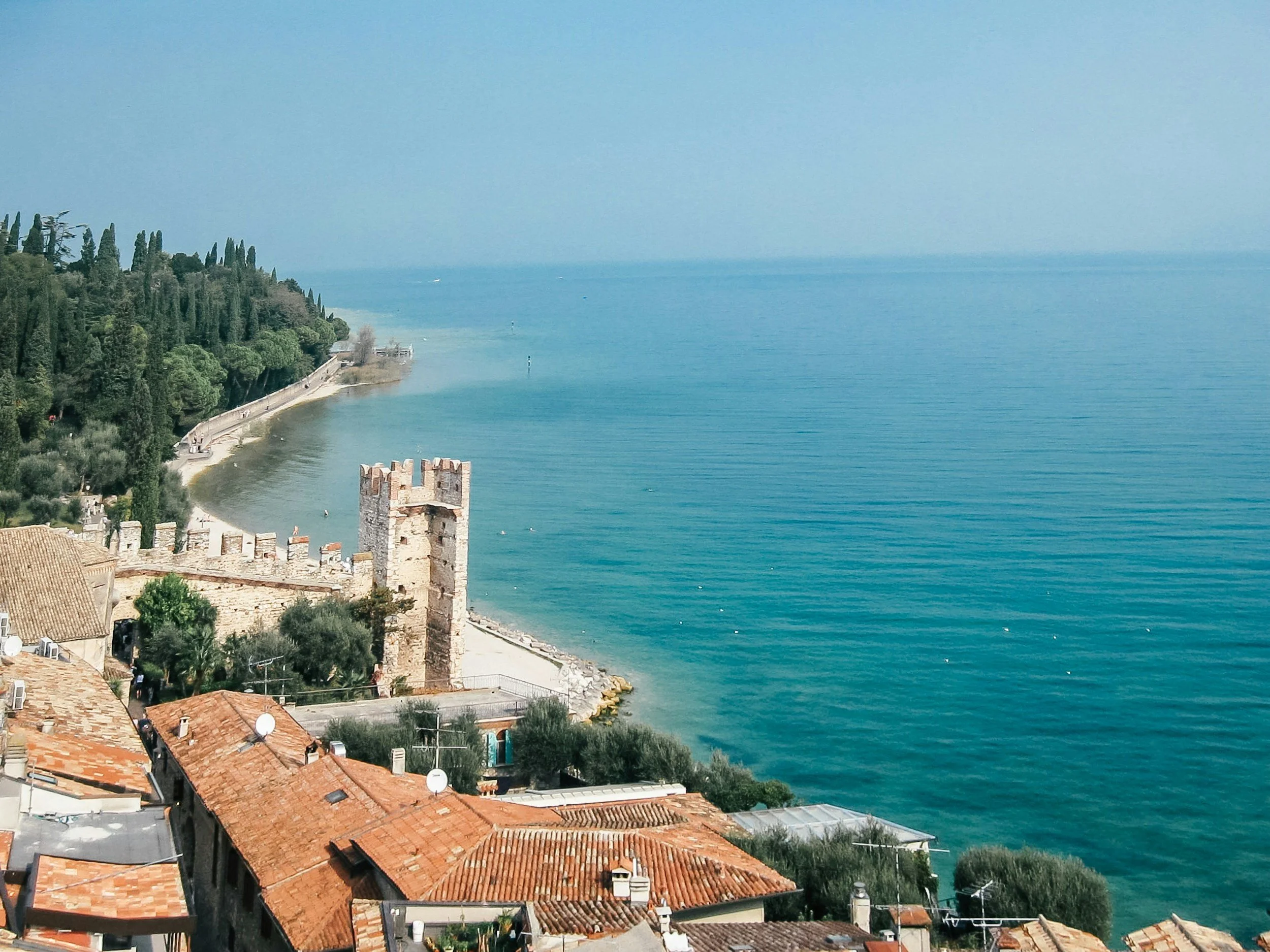 A scenic coastal view featuring red-tiled roofs of buildings, a medieval stone tower, a tree-lined shoreline, and a calm blue sea under a clear sky.