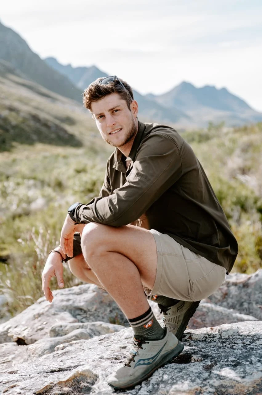 A man in hiking clothes squatting on a rocky terrain outdoors with mountains and greenery in the background.