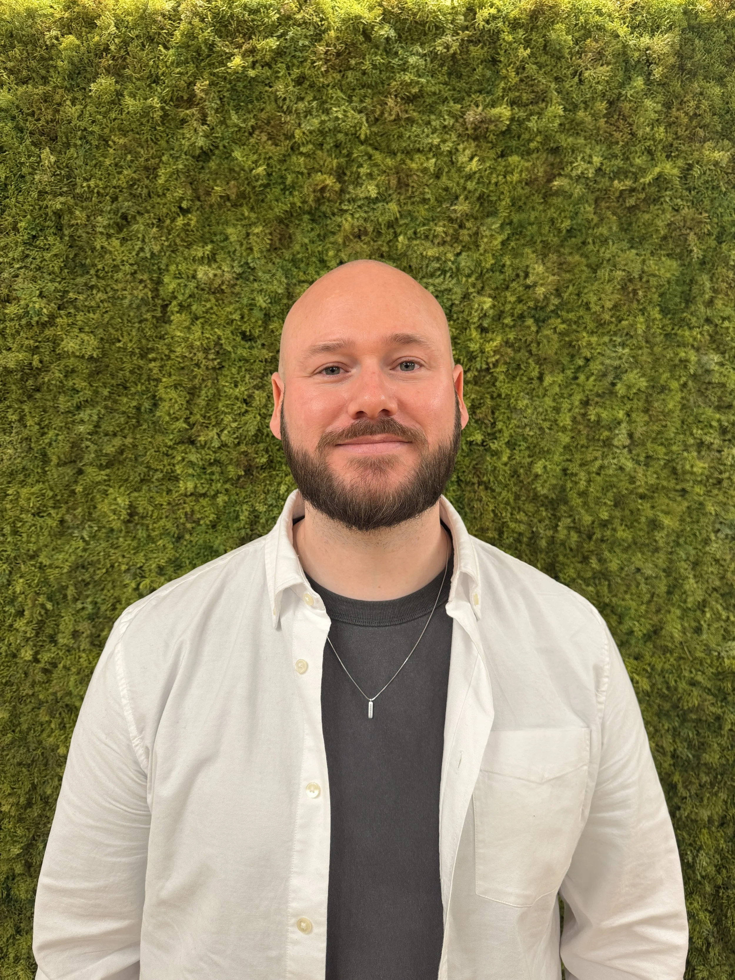 A man with a beard and a shaved head standing in front of a green mossy wall, wearing a white button-up shirt over a black t-shirt, with a silver necklace.