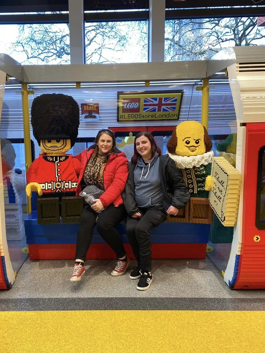 Two women sitting on a LEGO display of British characters outside a LEGO store. One is wearing a red jacket and the other a black jacket.