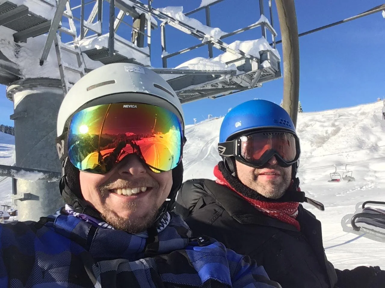 Two men wearing ski helmets and goggles sitting on a ski lift with snow-covered slopes and ski chairs in the background on a sunny day.