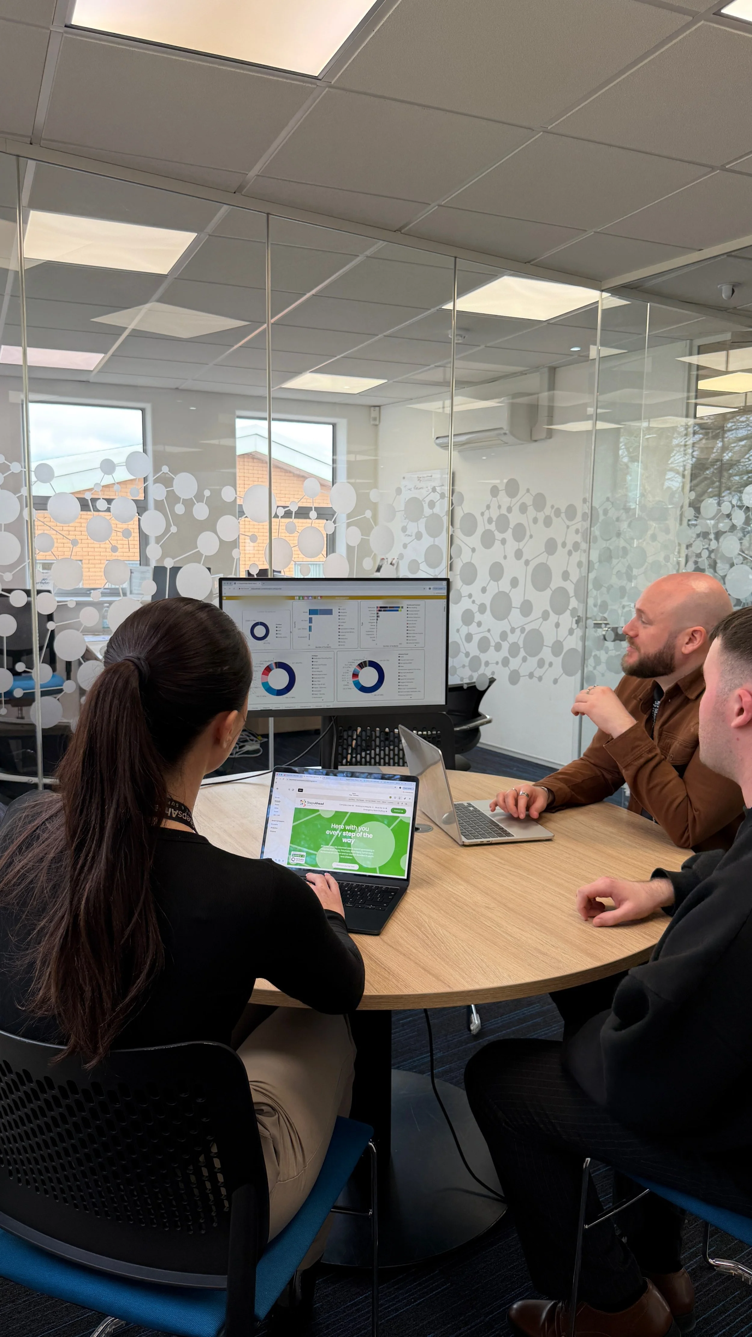 Three people sitting around a conference table in an office, discussing data on a monitor and a laptop, with glass walls and windows in the background.