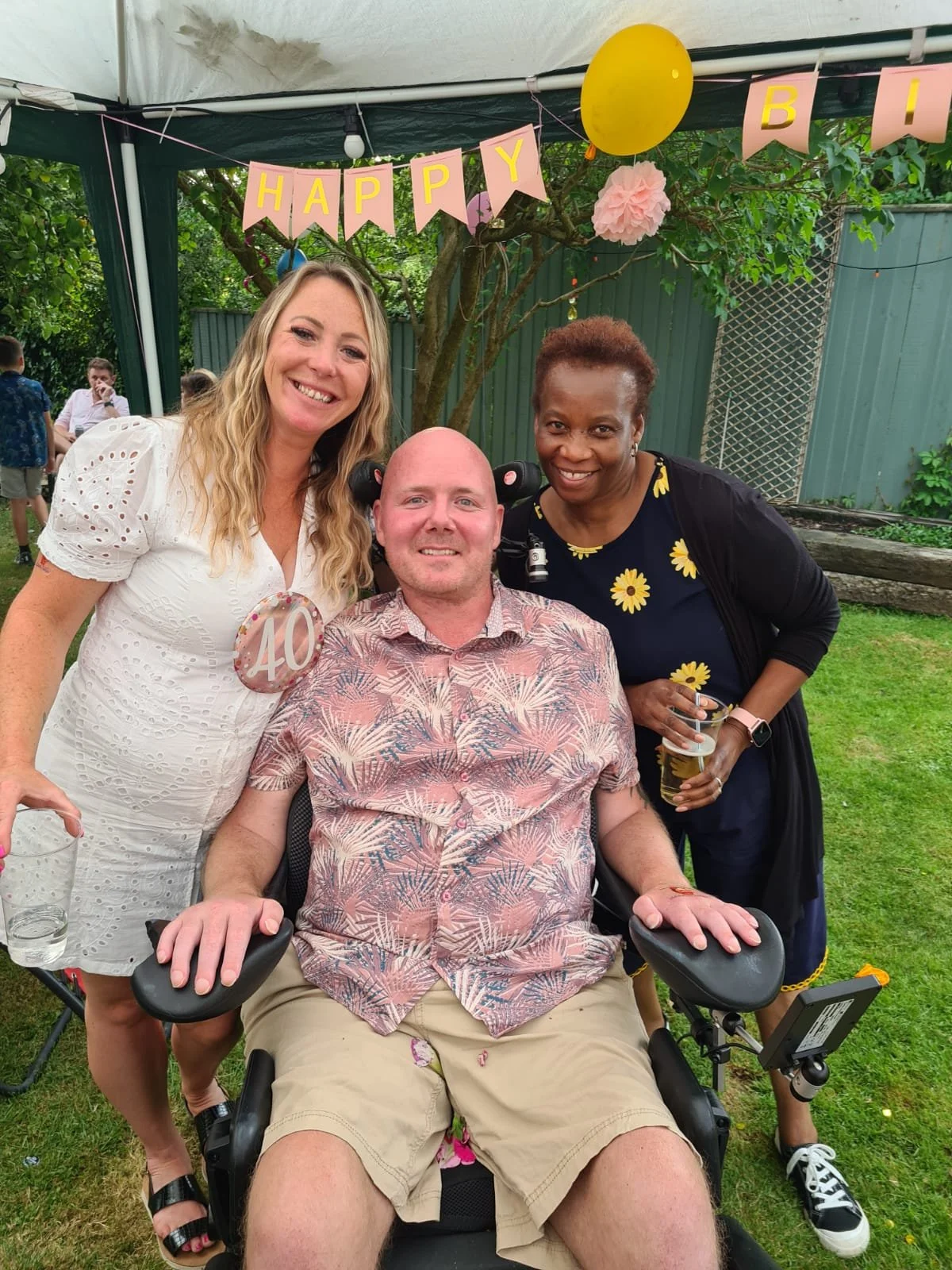 Three people at a birthday celebration, with a banner reading 'HAPPY BIRTHDAY' and a yellow balloon in a backyard garden. One person is in a wheelchair, and the others are standing beside him, smiling.