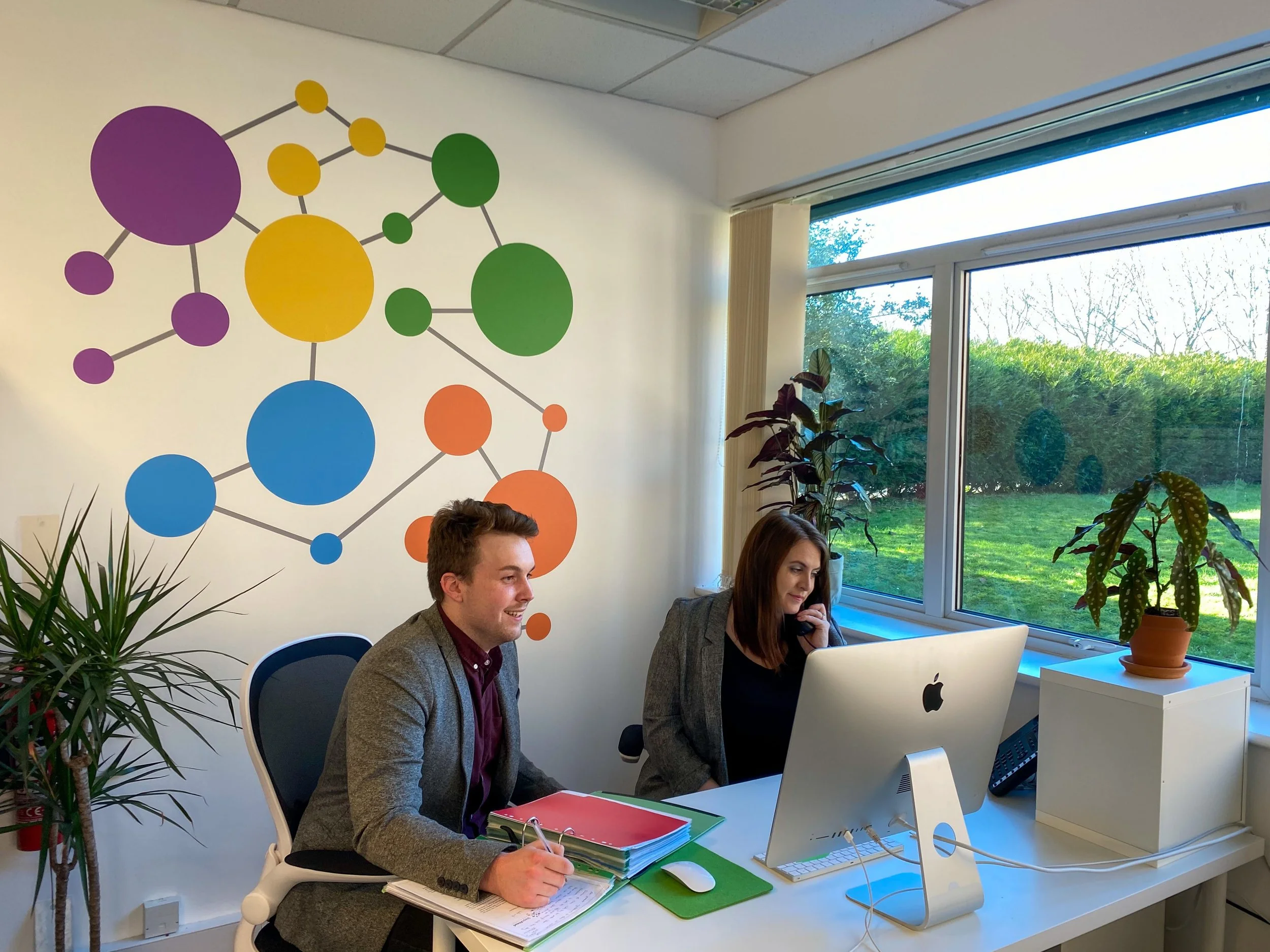 Two people sitting at a desk in an office, one woman talking on the phone and one man smiling with notebooks, white computer and potted plants, with large window showing green outdoor scenery, and colorful abstract wall art.