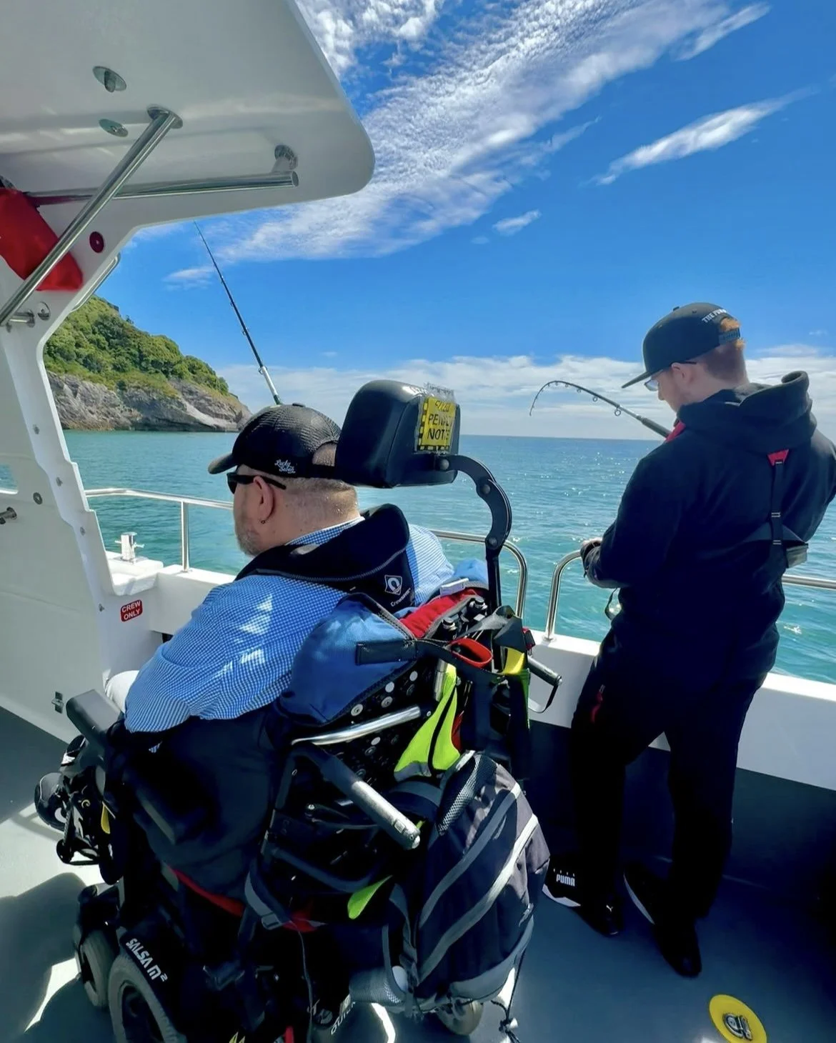 Two men on a boat, one in a wheelchair, fishing in a sunny blue ocean near a rocky shoreline covered in green trees.
