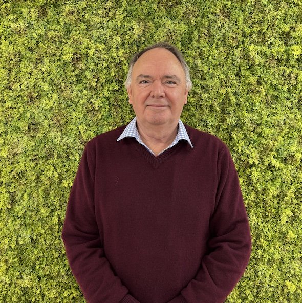 A middle-aged man with gray hair and a slight smile, wearing a maroon sweater over a collared shirt, standing in front of a green leafy wall.