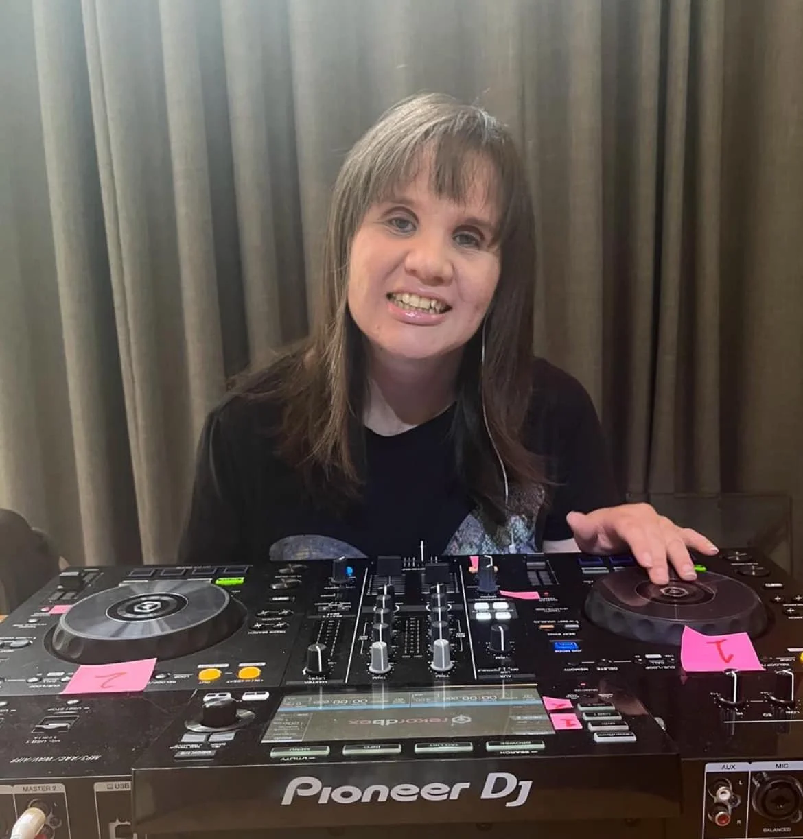 Woman with long brown hair smiling behind a Pioneer DJ controller, wearing headphones, with pink sticky notes on the equipment and gray curtains in the background.