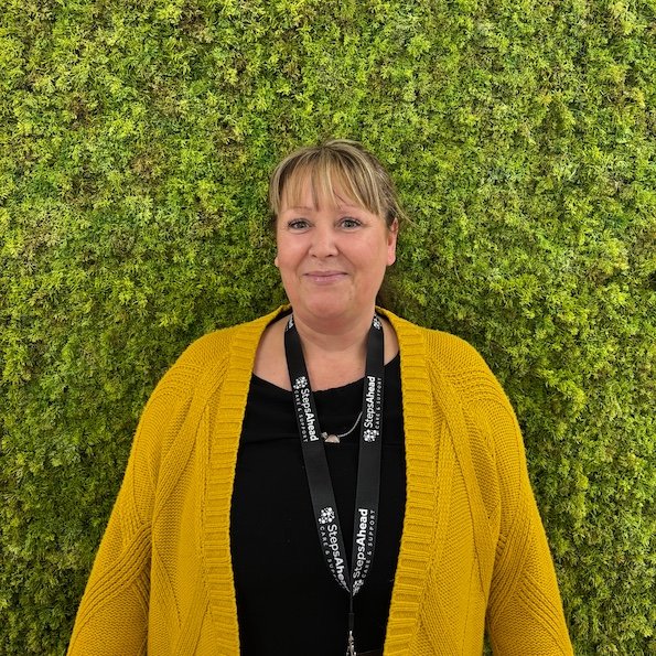A woman with short blonde hair and a black lanyard, wearing a yellow cardigan over a black shirt, standing in front of a green moss wall.