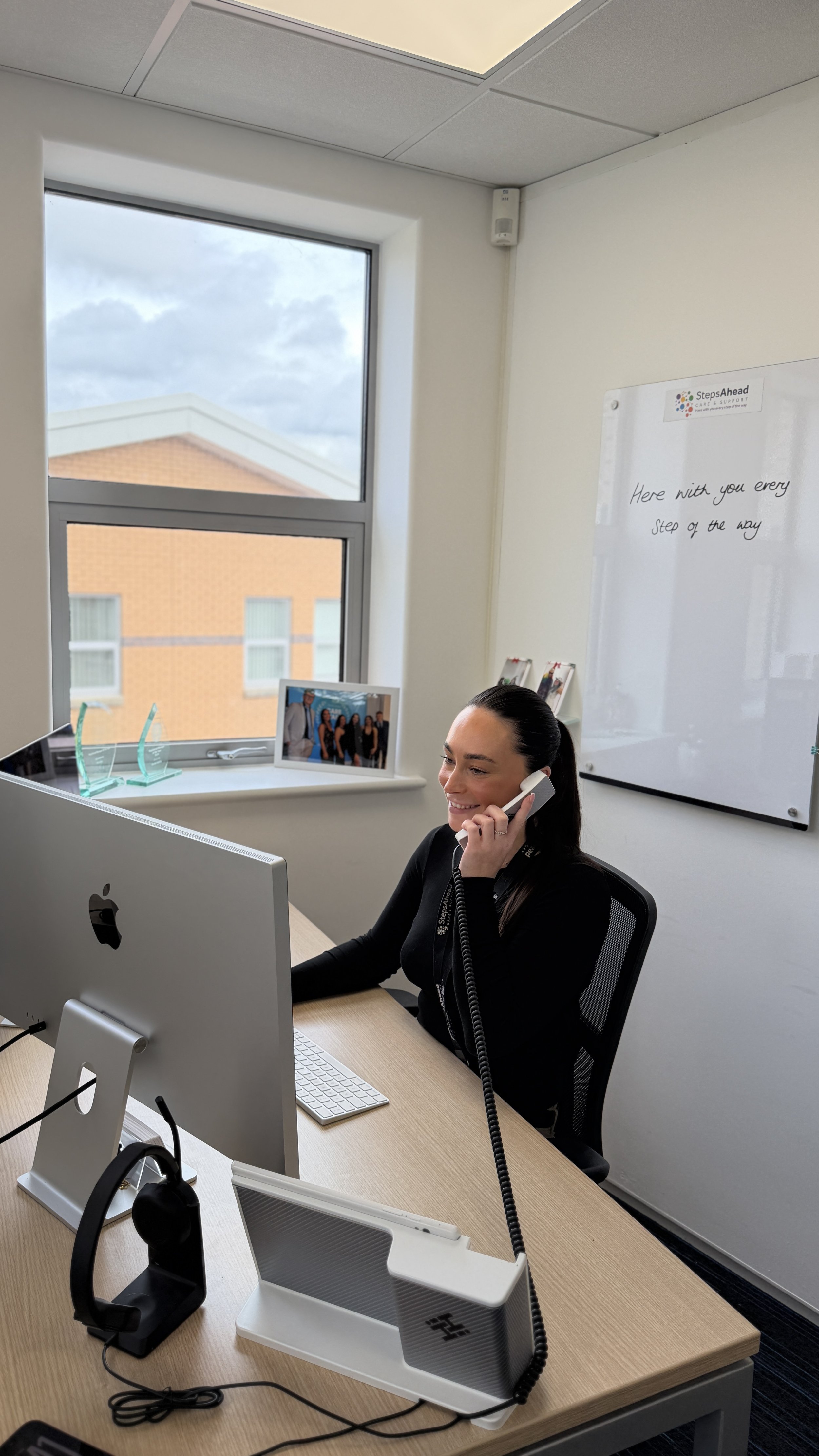 Woman working at a desk in an office, talking on the phone and smiling, with a large monitor, keyboard, and framed photo on the windowsill, and a whiteboard on the wall.